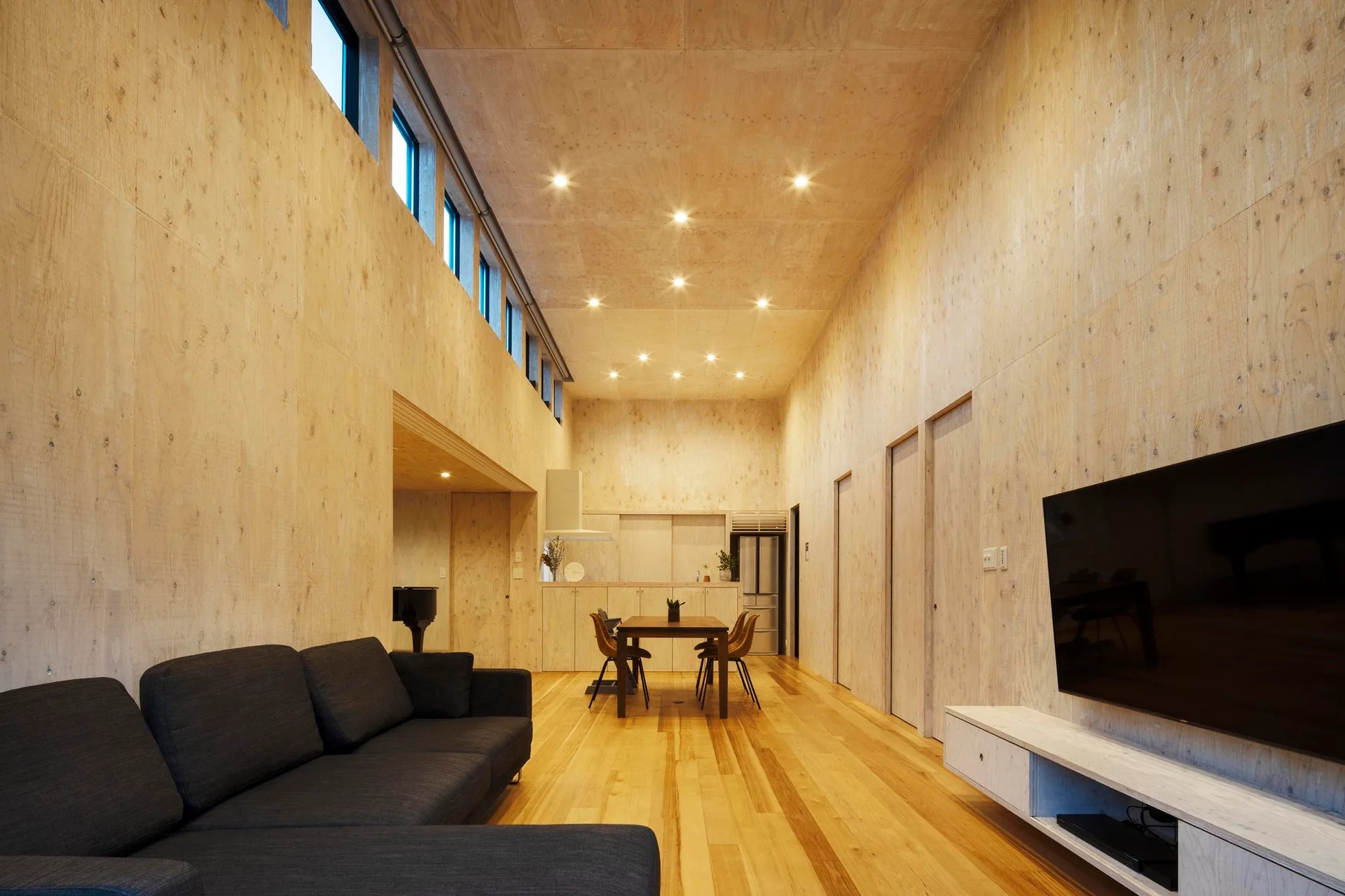 A minimalist living and dining space with plywood paneled walls and ceiling, a dark grey sectional sofa, and a row of high clerestory windows.