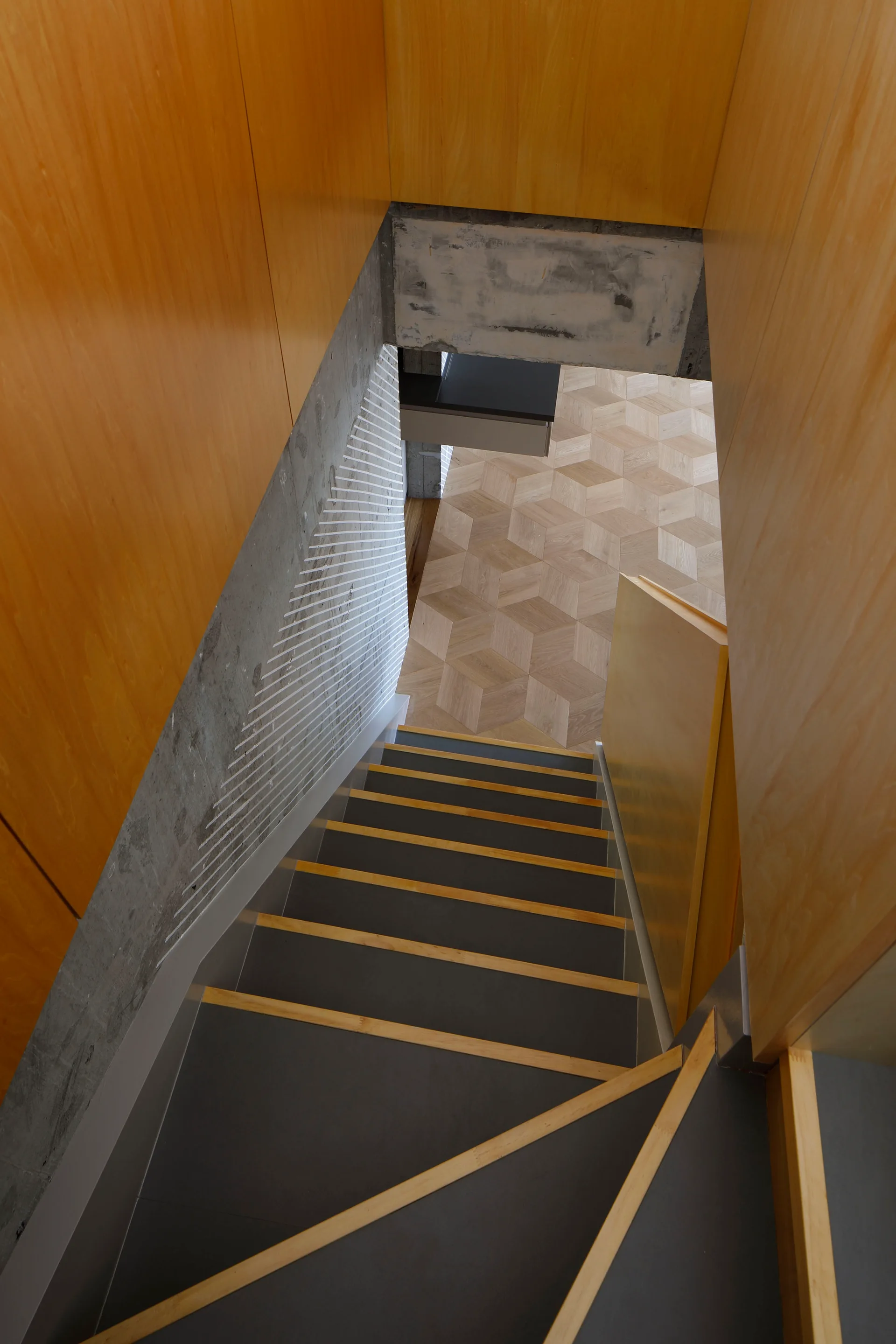 A high-angle view looking down a modern staircase with dark treads and light wood edges, flanked by plywood walls and a concrete support.