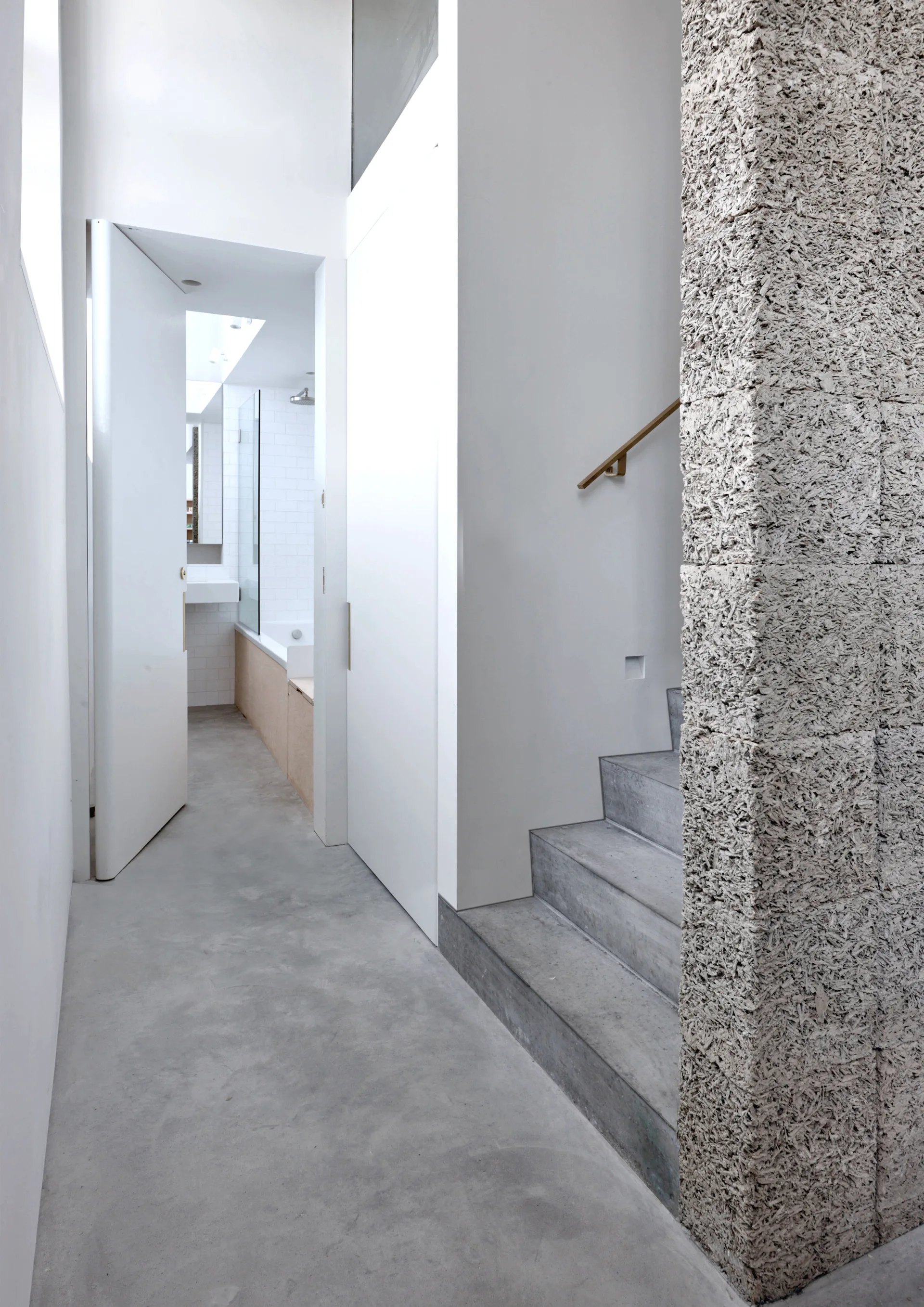 A minimalist hallway featuring polished concrete floors, a concrete staircase, and a rough-textured grey column leading past a white bathroom doorway.