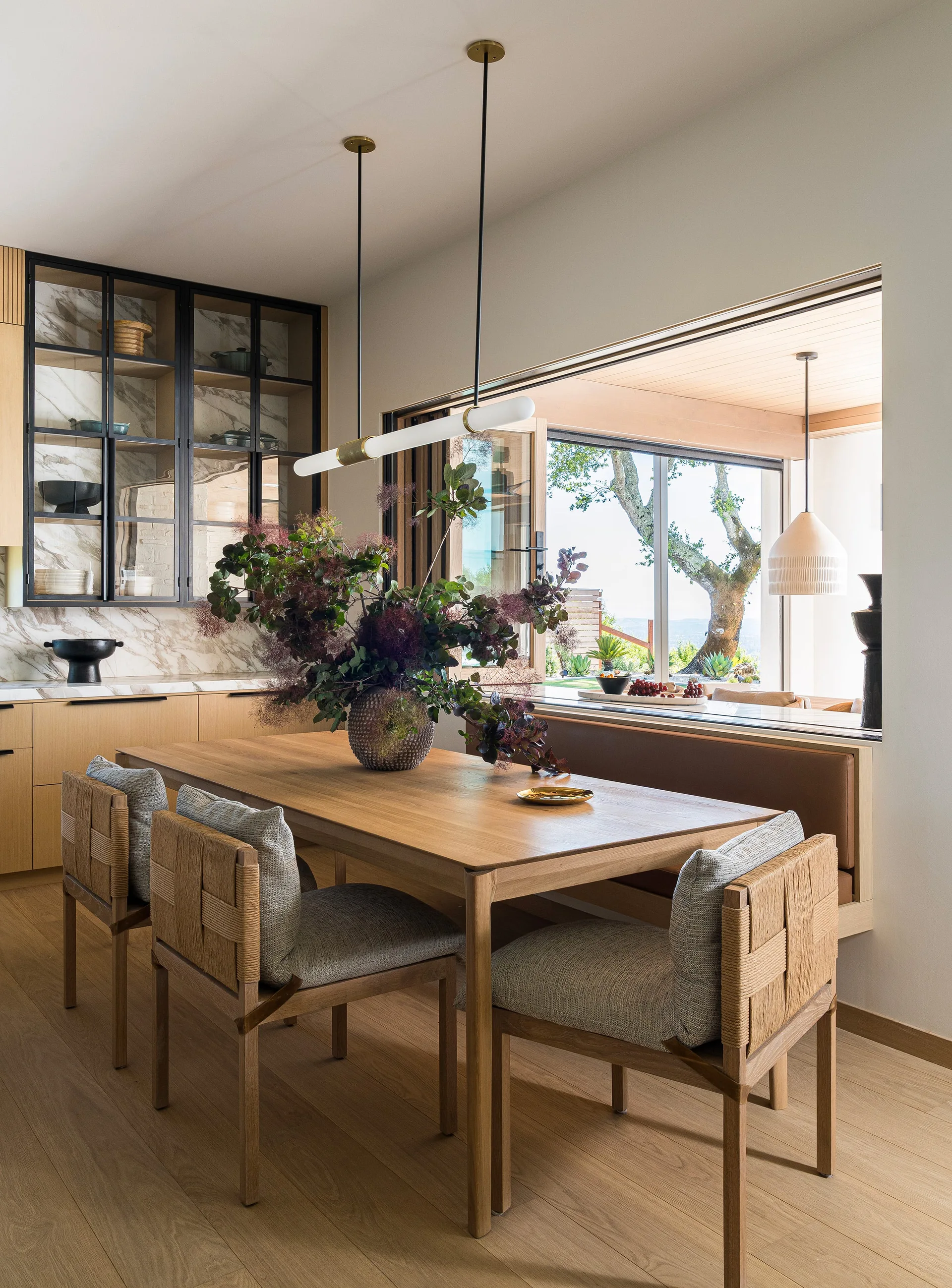 A wooden dining table is surrounded by woven back chairs and a leather-upholstered built-in banquette under a large open window pass-through.
