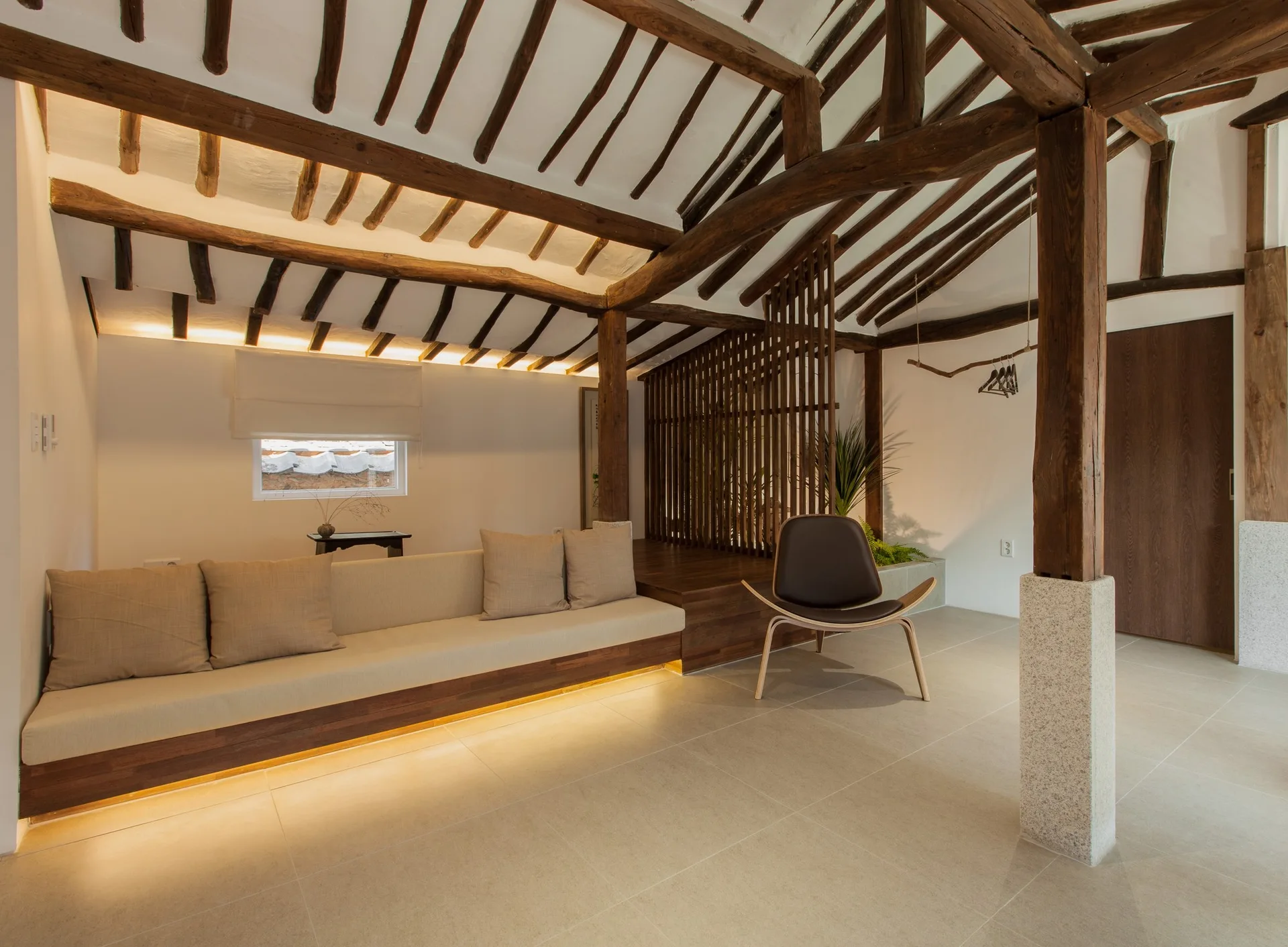 A minimalist lounge features a low built-in platform sofa, exposed dark wood ceiling rafters, a slatted wooden screen, and large tile floors.