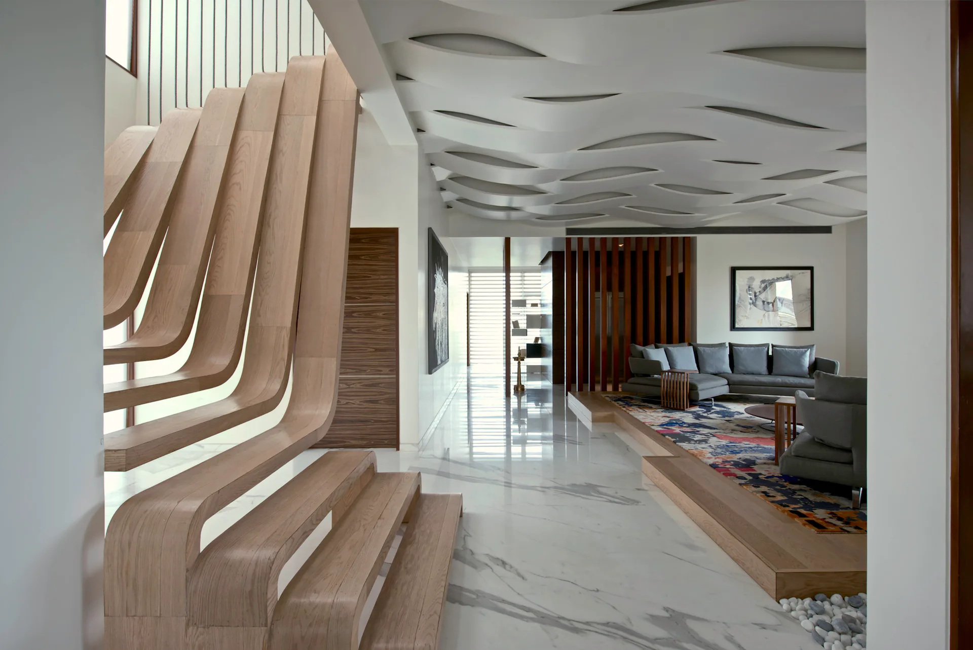 A modern interior features a sculptural ribbon-like wooden staircase, a wavy plaster ceiling, and a grey sectional sofa on a raised platform.