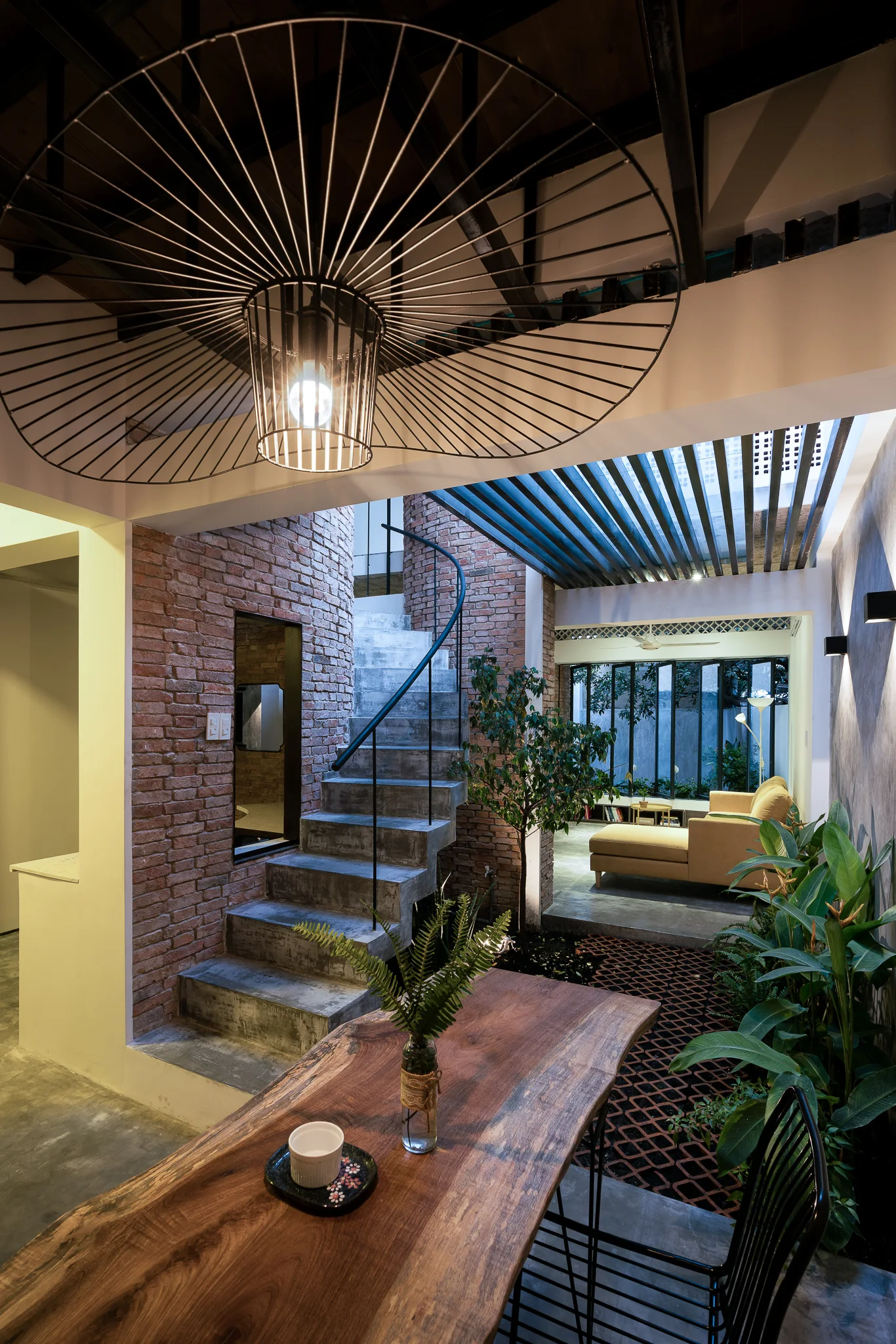 A modern open-concept home interior featuring a concrete staircase against brick walls next to a rustic wood dining table with a black wire pendant light.