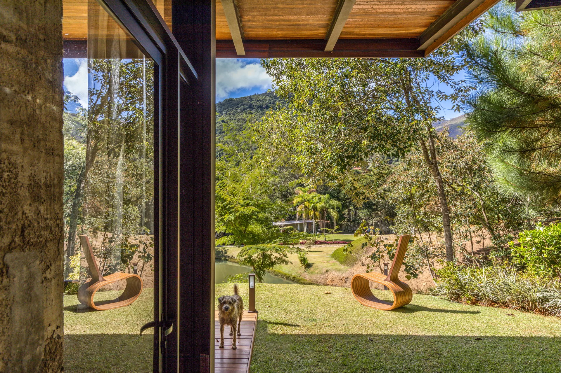 An open glass door looks out onto a wood patio deck and green lawn with two sculptural curved wood lounge chairs and a scenic mountain view.