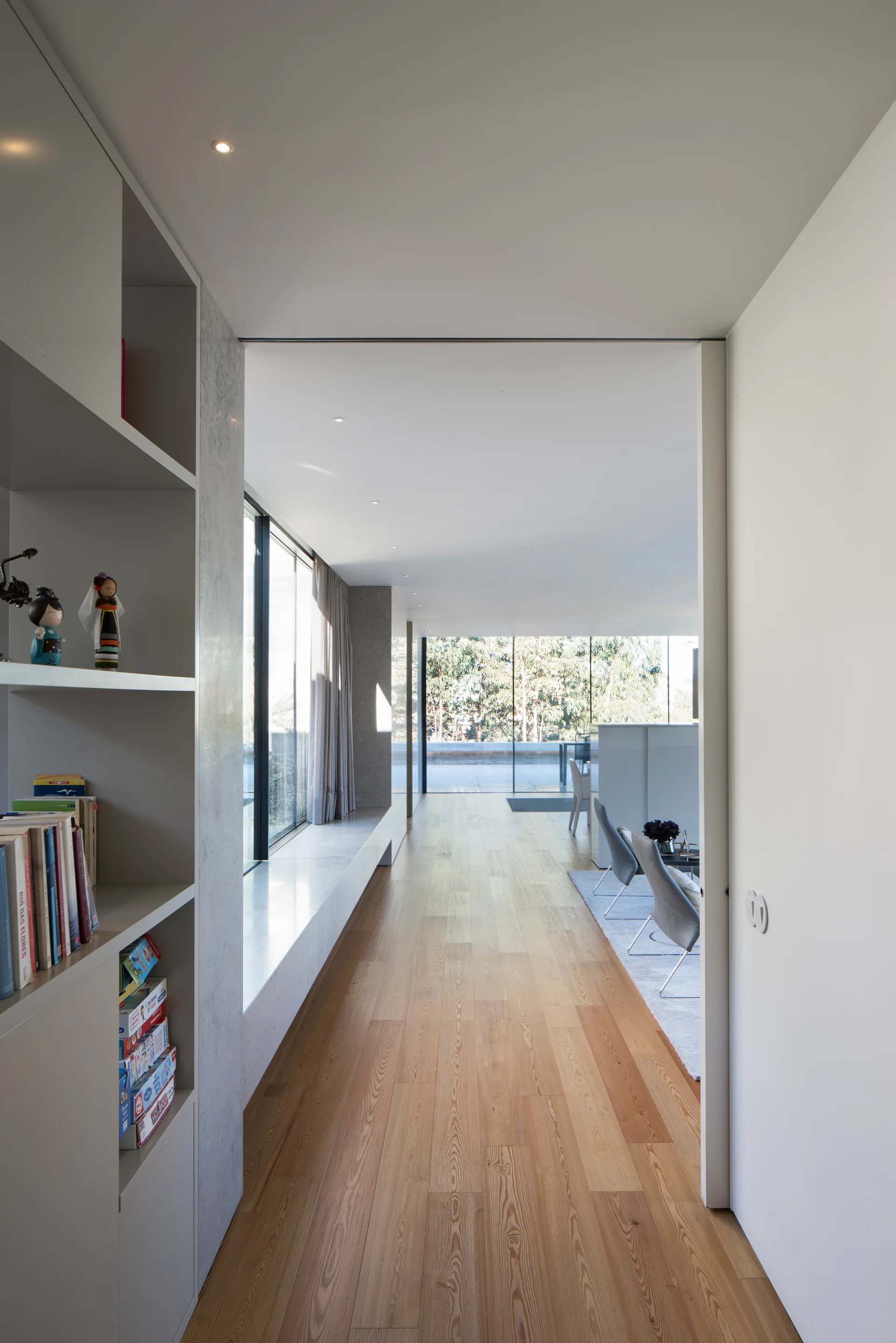 A long hallway with light wood flooring features built-in white shelving on the left and leads to a bright living room with large glass windows.