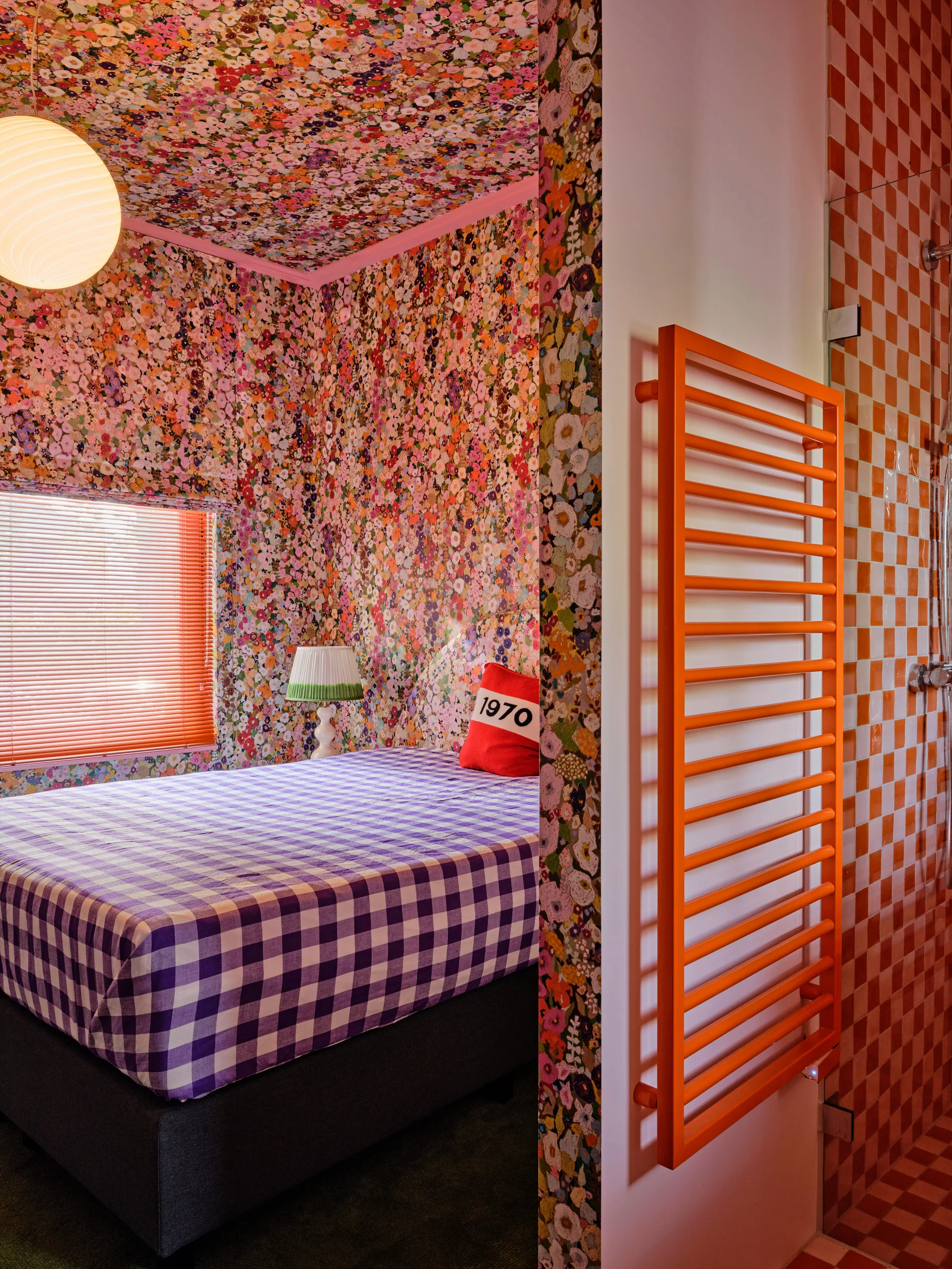 A bedroom with dense floral wallpaper on the walls and ceiling, purple gingham bedding, and a view into an orange tiled bathroom.