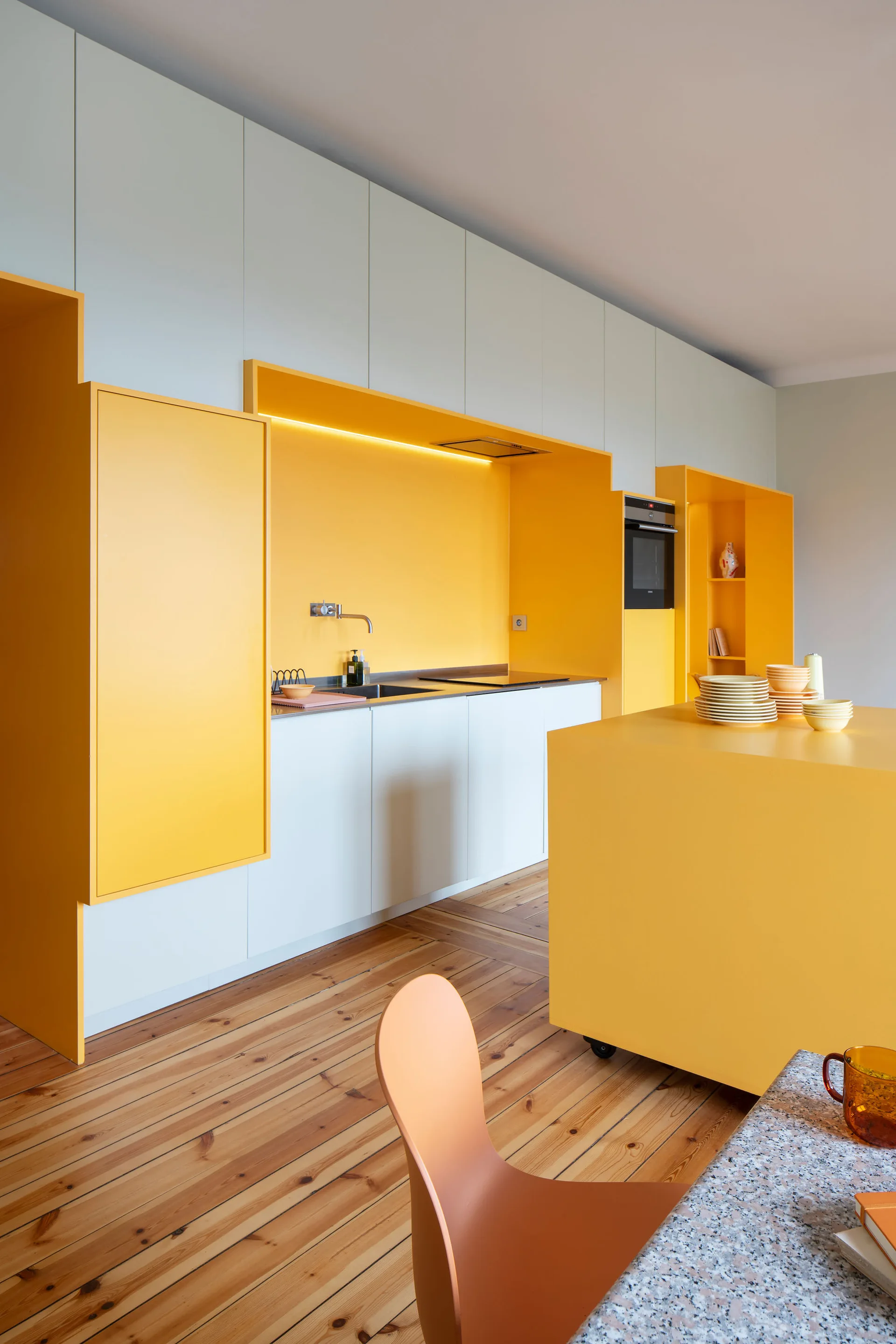 A minimalist kitchen features bold yellow and white cabinetry, a yellow rolling island, a metal faucet, and natural wood plank flooring.