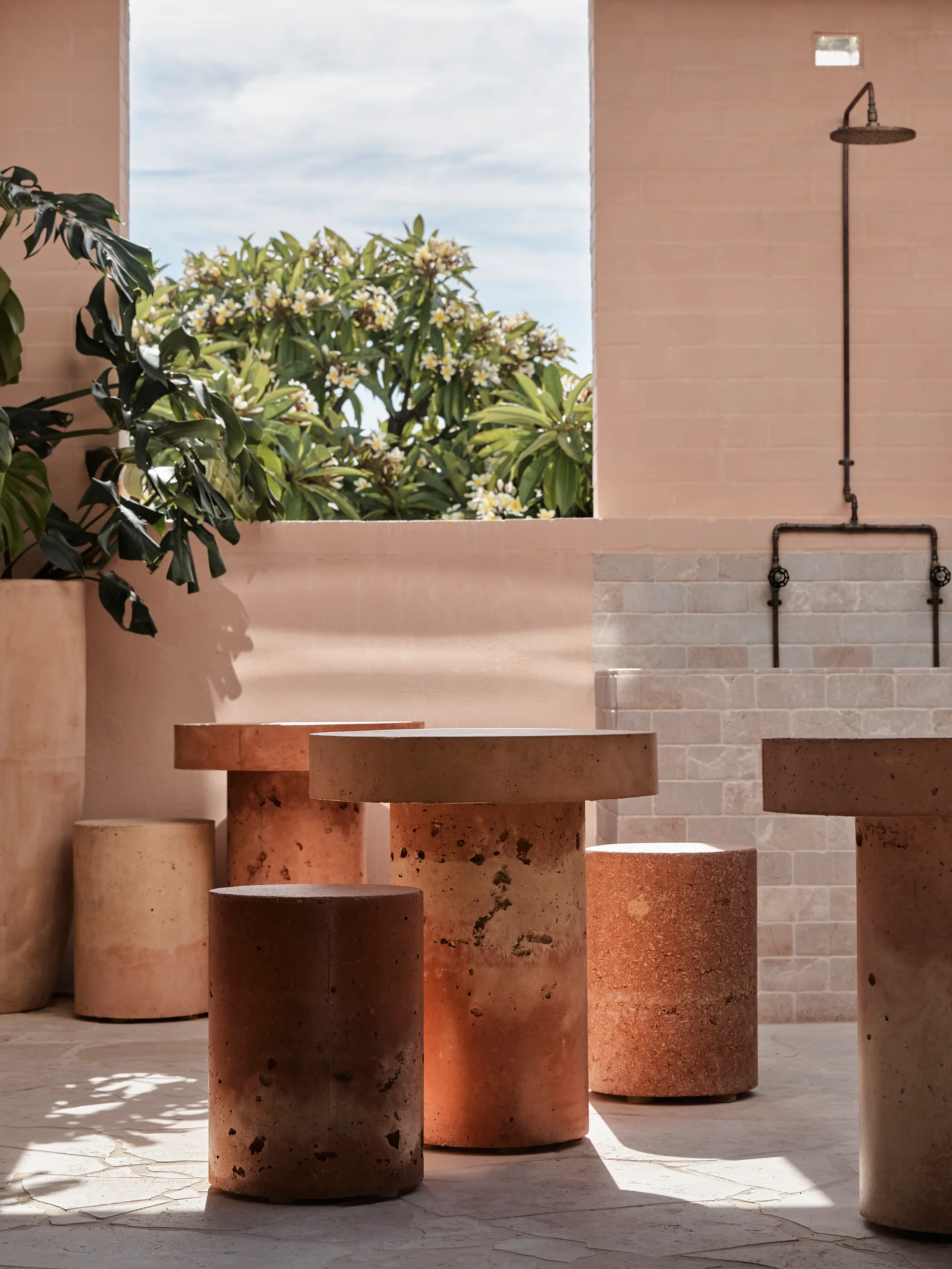 An outdoor patio features peach brick walls, an integrated outdoor shower, and cylindrical stone tables and stools on an irregular paved stone floor.