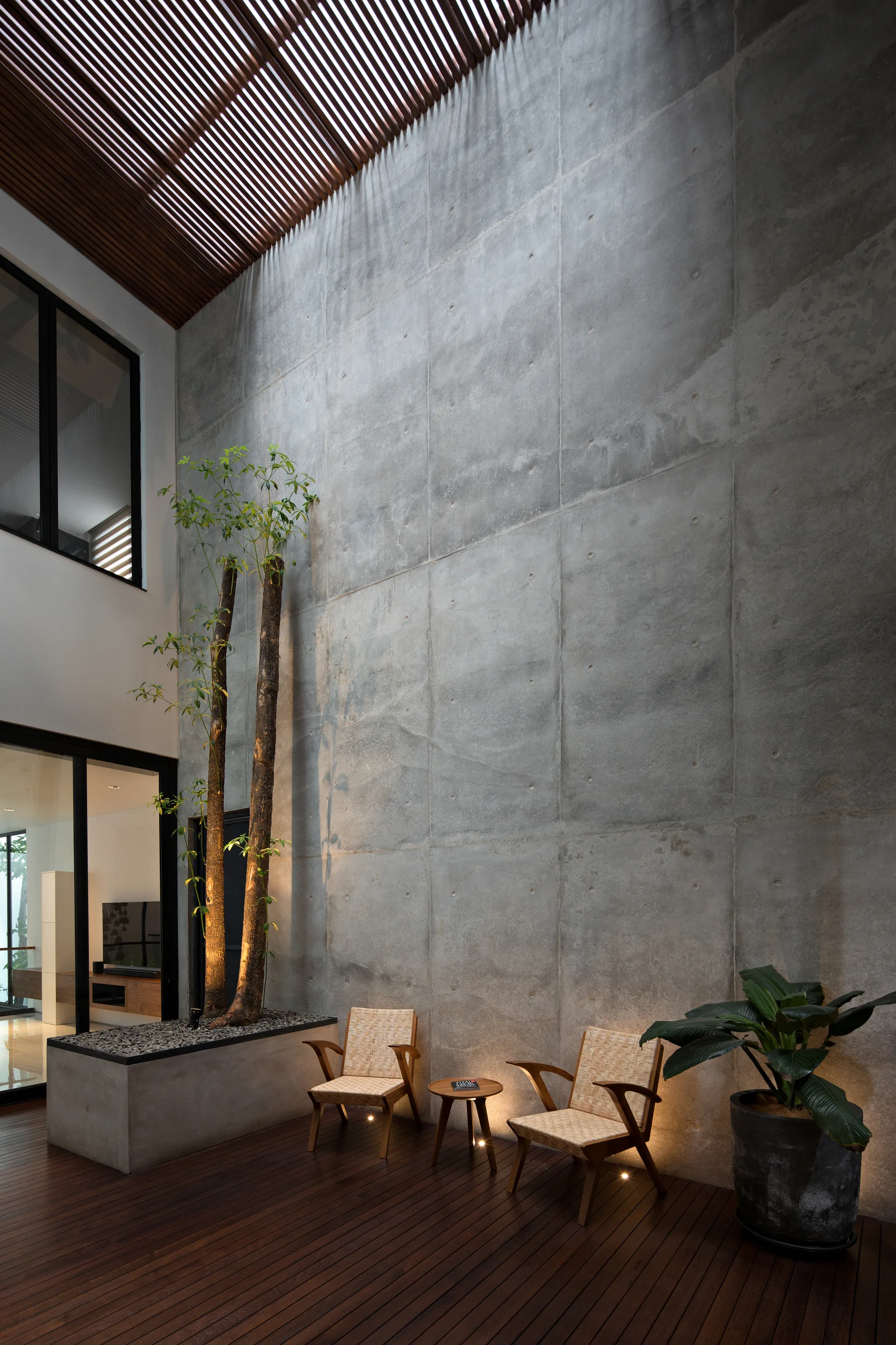 A double-height indoor atrium features a towering concrete wall, dark wood decking, a slatted timber ceiling, and two woven lounge chairs.