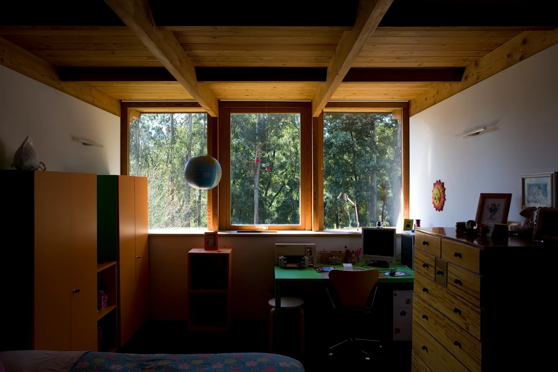 A children's bedroom features a wood beam ceiling, a green desk under a large window looking at trees, orange wardrobes, and a wooden dresser.