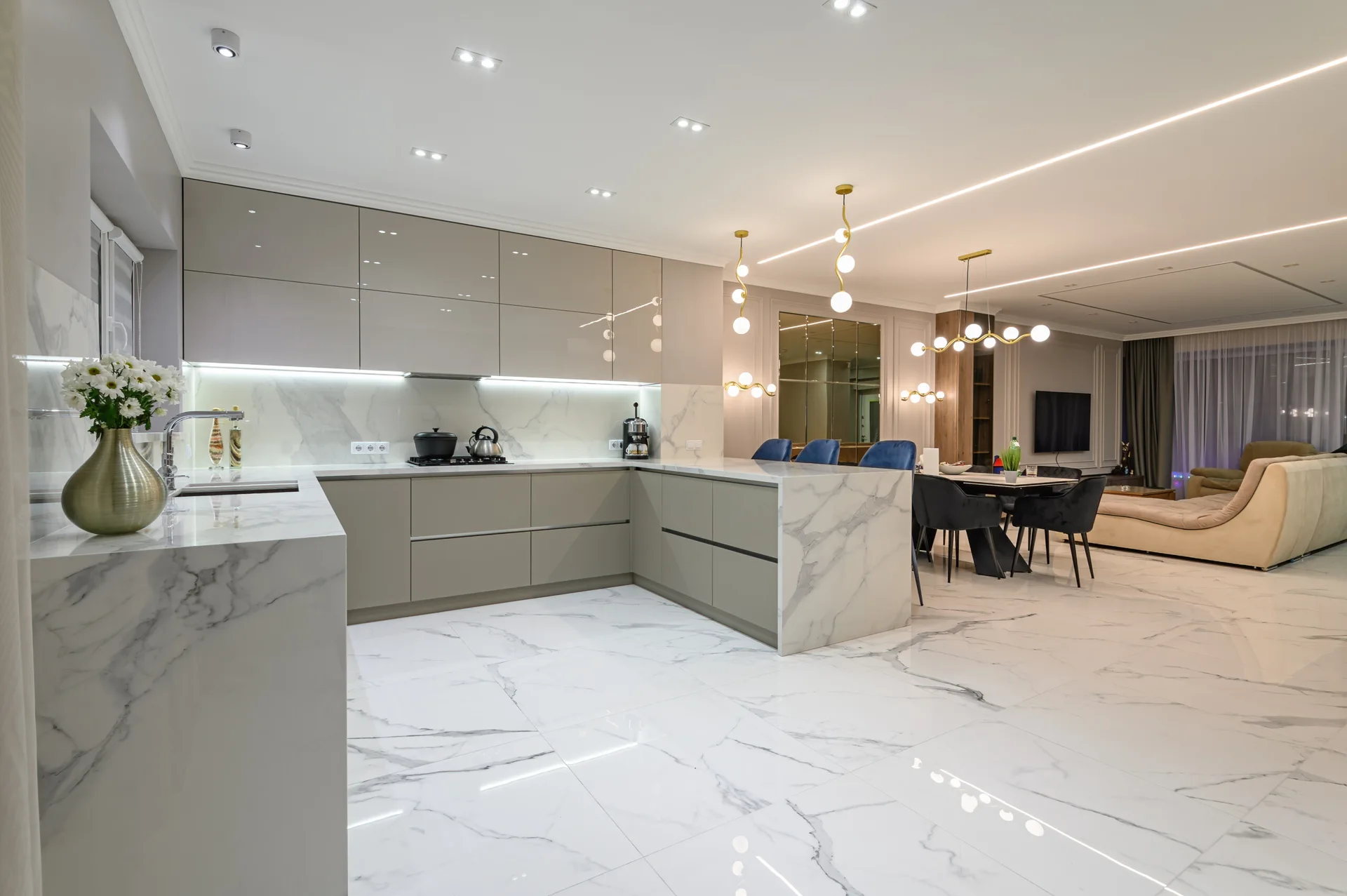 A modern open-plan kitchen with glossy grey cabinetry, marble-patterned flooring and countertops, and a dining area visible in the background.
