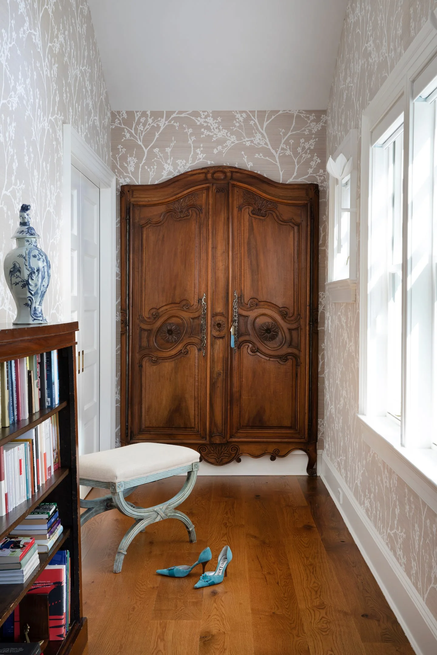 A traditional hallway features a large ornate wooden armoire, botanical patterned wallpaper, a small upholstered bench, and hardwood flooring.