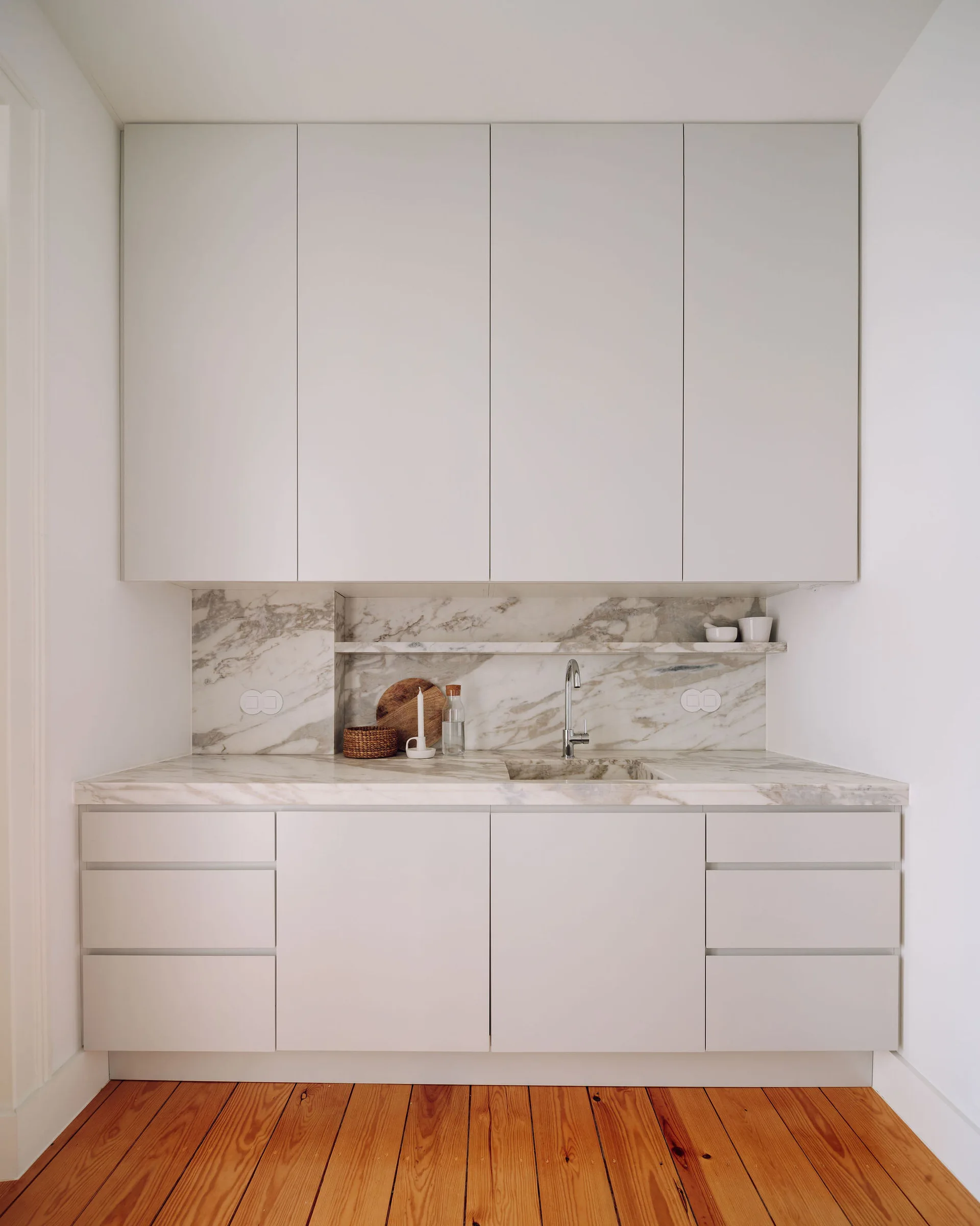 A minimalist kitchen alcove with light gray handleless cabinets, a heavily veined marble countertop, an integrated marble shelf, and natural wood floors.