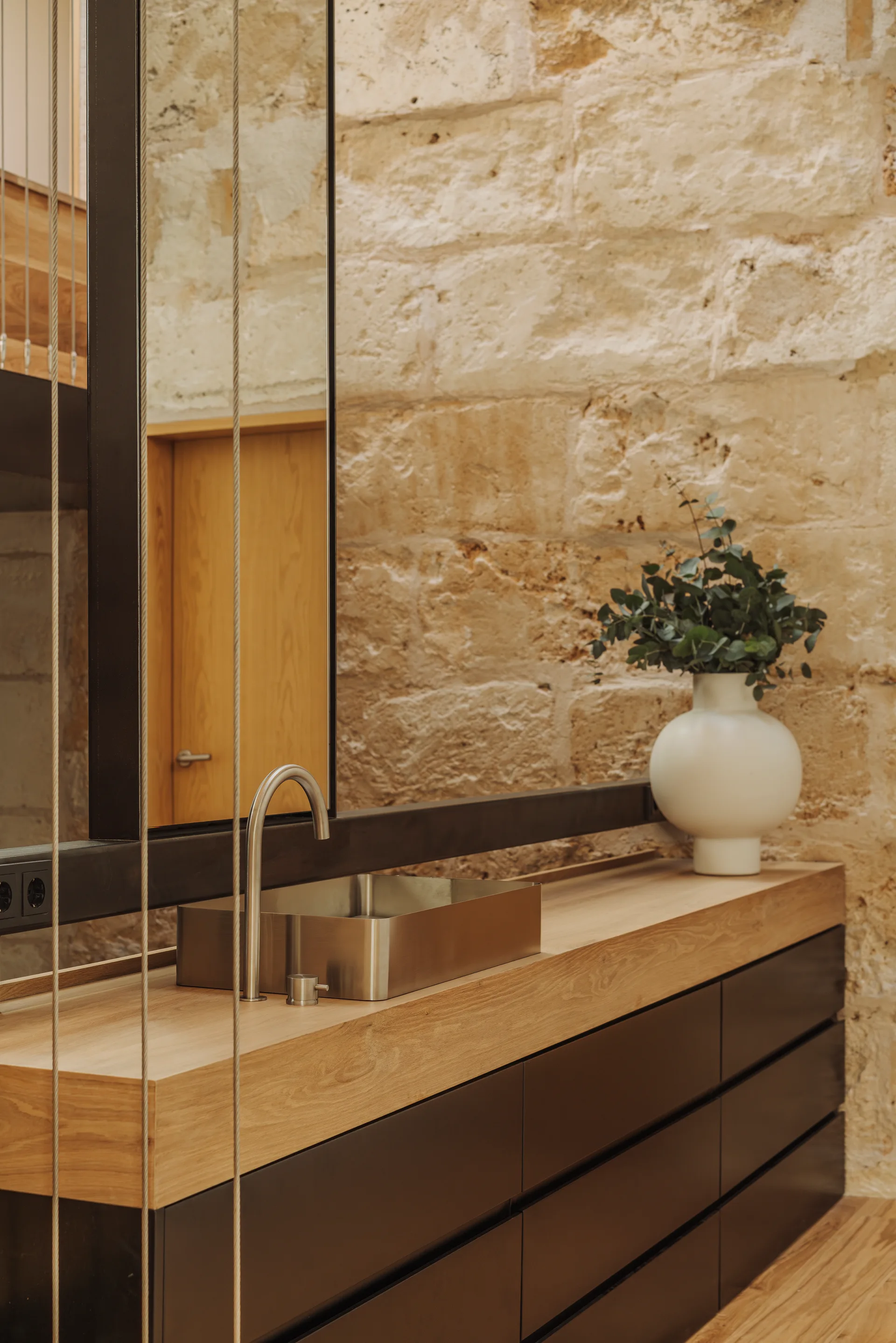 A modern bathroom vanity featuring a wooden countertop, a stainless steel rectangular sink, and dark lower drawers set against a rough stone wall.