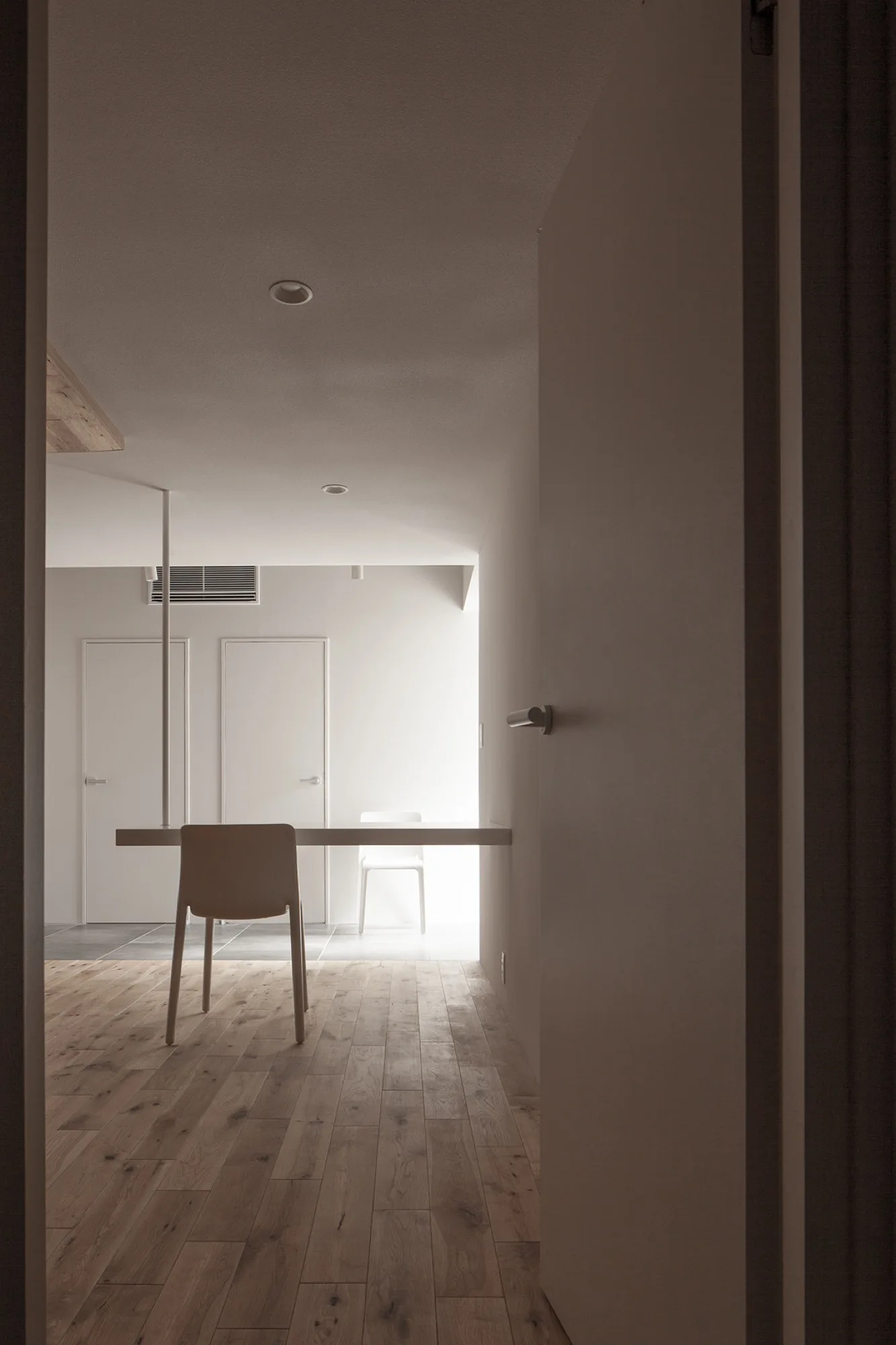 An interior view through a doorway shows a minimalist dining table and chair on light wood plank flooring against white walls.