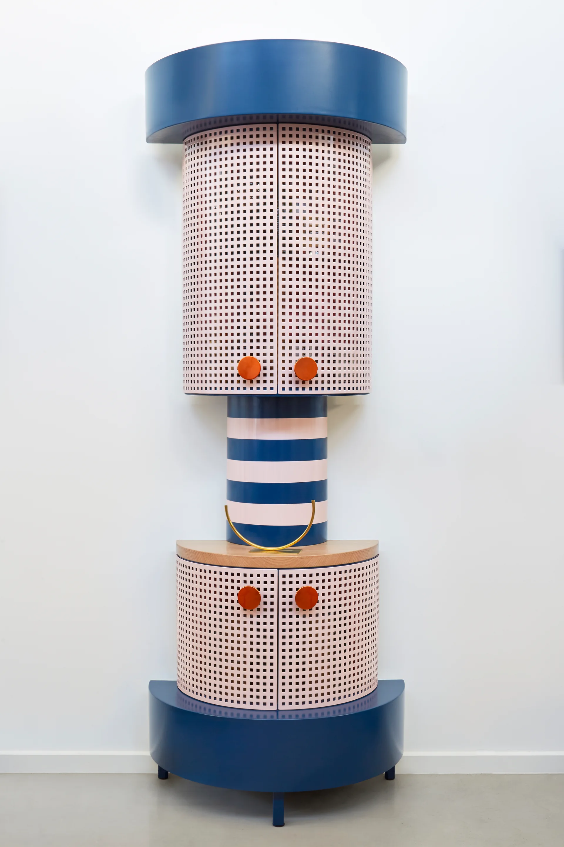 A tall, cylindrical storage cabinet with pink perforated doors, navy blue cap and base, and a striped middle section stands against a white wall.