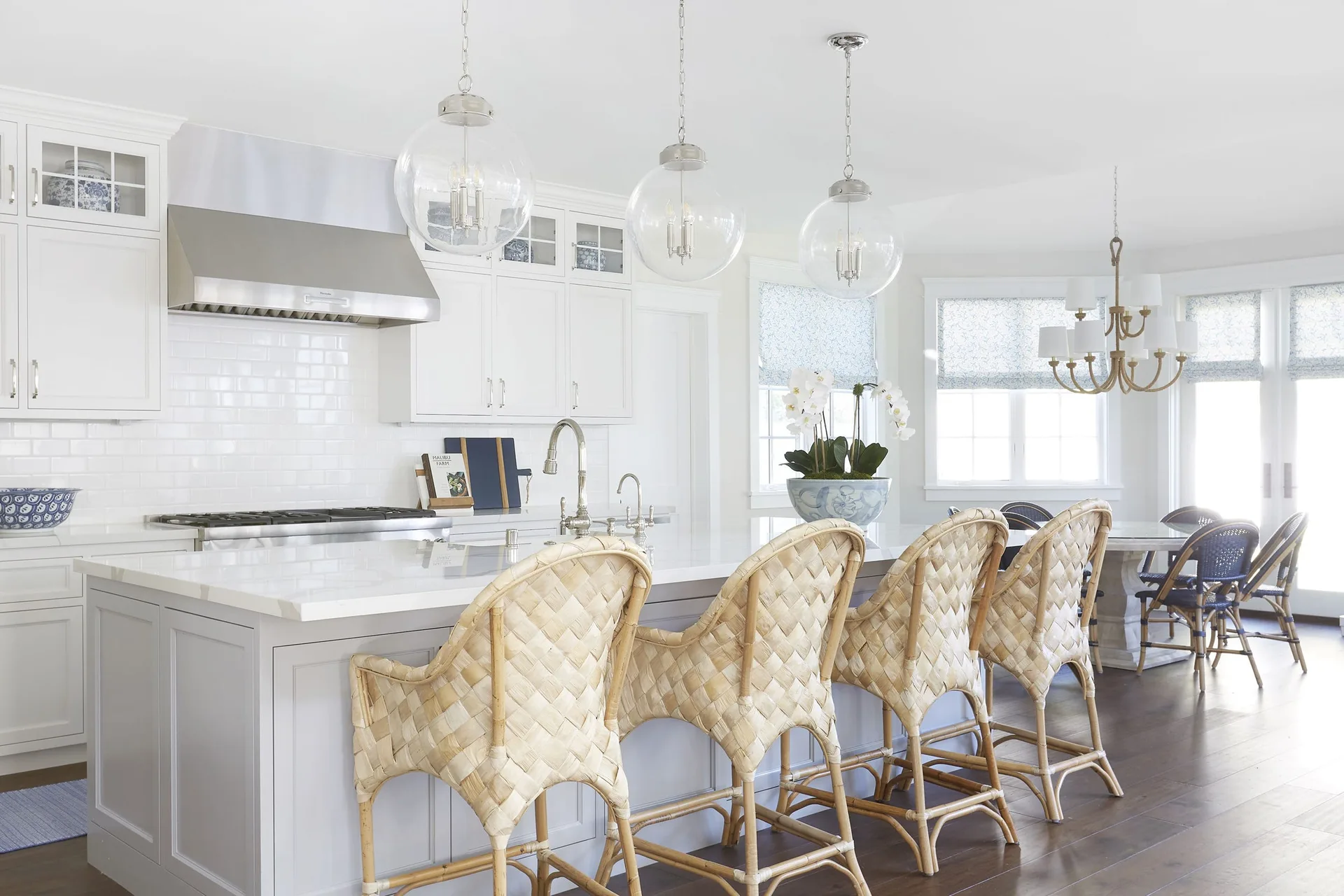 A bright white kitchen features a large island with four woven bar stools, glass globe pendant lights, and a view of a dining area with blue chairs.