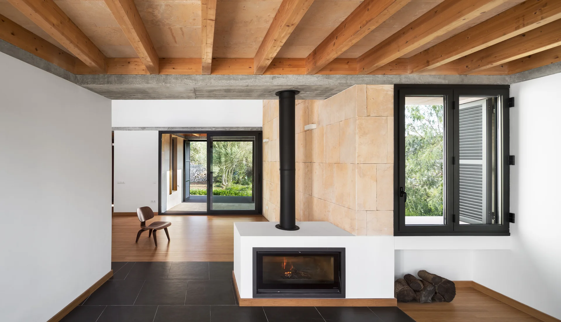 A contemporary living room featuring a minimalist white fireplace with a black chimney pipe, a stone accent wall, and exposed wooden ceiling beams.