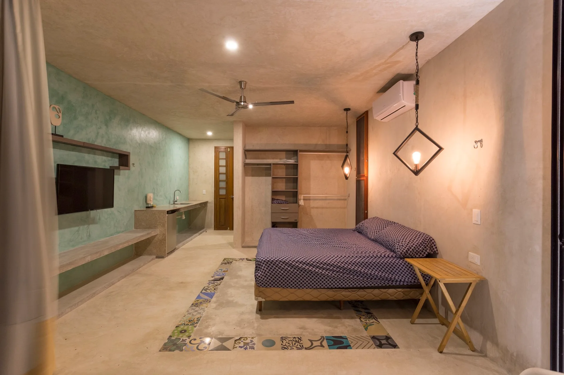 A studio bedroom featuring concrete-finished walls and flooring, a bed with blue patterned bedding, a patterned tile border inlay, and a teal accent wall.