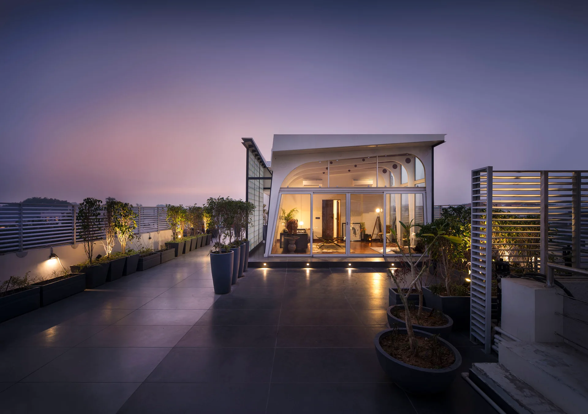 A wide dark grey tiled rooftop terrace features a glass pavilion lit from within, surrounded by dark planters with uplighting at twilight.