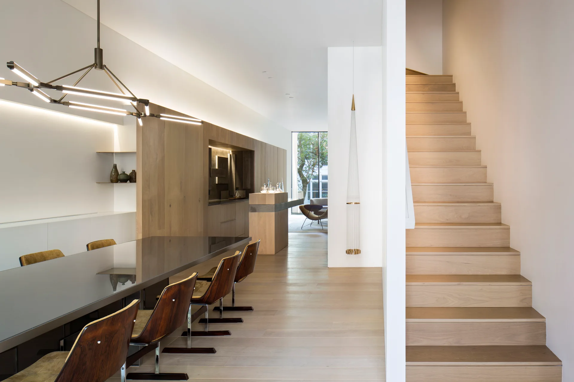 A modern dining room featuring a dark reflective dining table, mid-century wooden chairs, light oak wall paneling, and an adjacent wooden staircase.