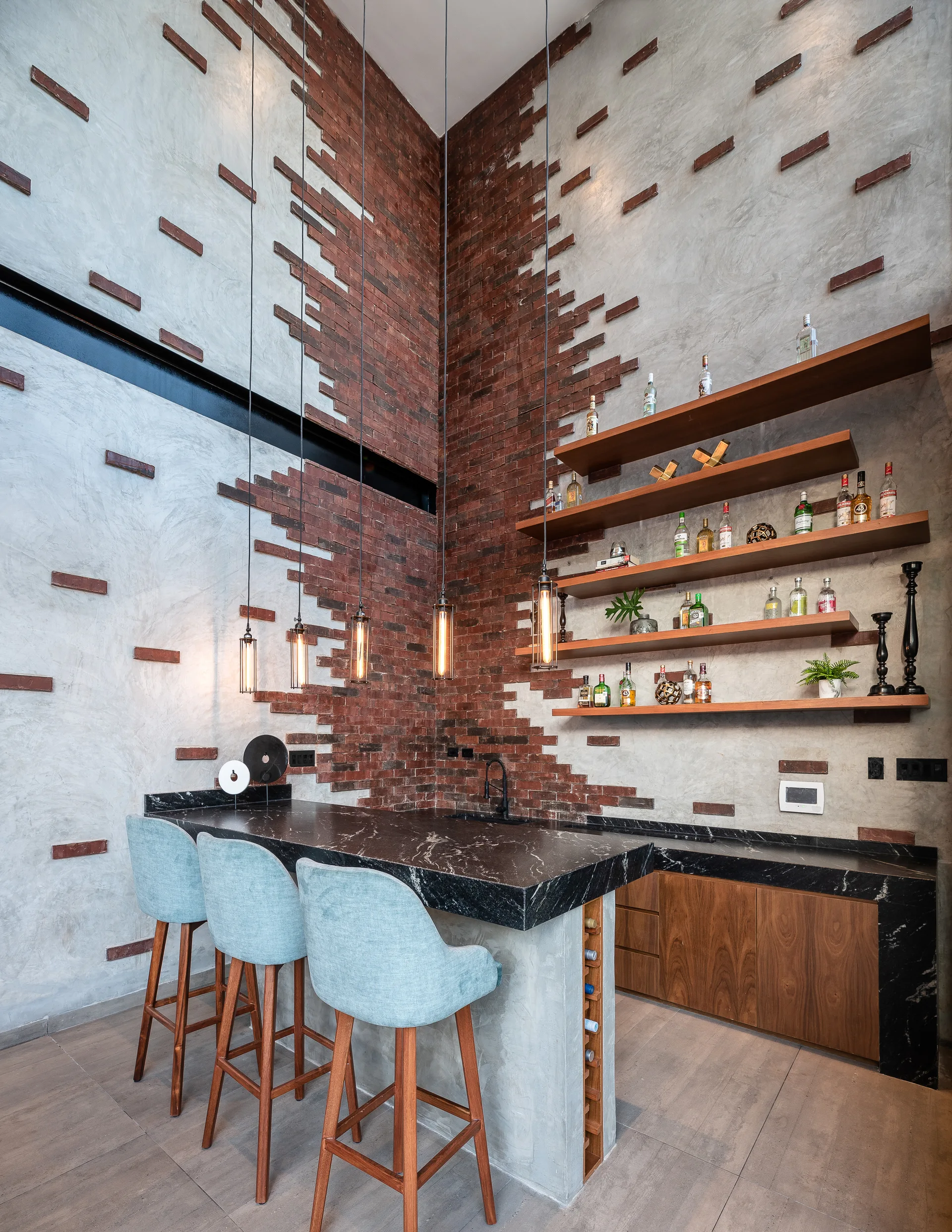 A home bar features blue upholstered stools at a dark stone-topped concrete island in front of an exposed brick and plaster wall.