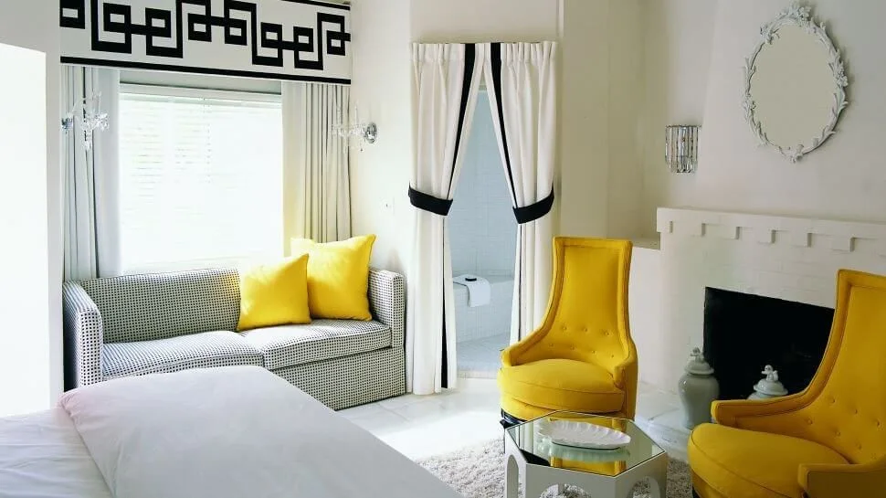 A bedroom sitting area features two yellow high-back armchairs, a patterned sofa, a white brick fireplace, and drapes with black trim.