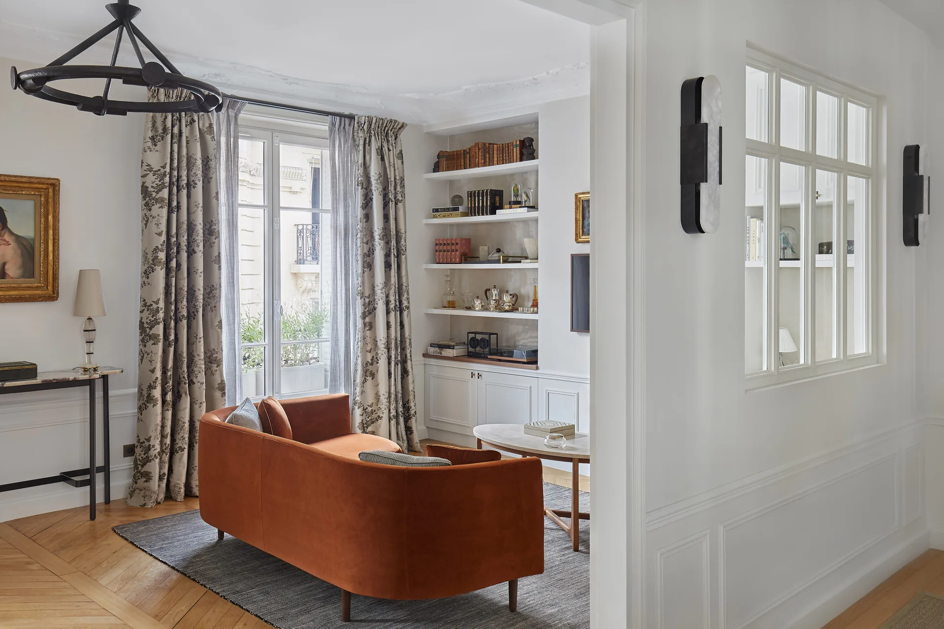 A living room features a curved orange sofa, an oval coffee table, built-in shelving, and large windows with patterned curtains.