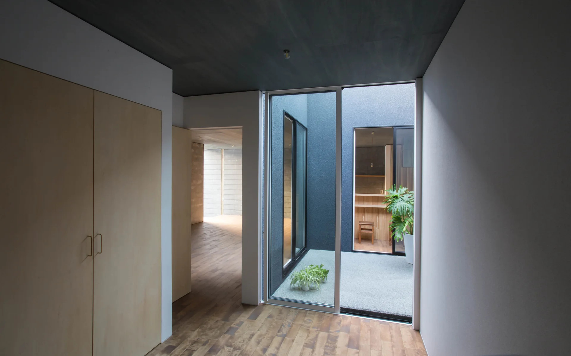 A minimalist hallway features a light wood wardrobe, wood plank floors, and a dark grey ceiling next to a glass door opening to a gravel courtyard.