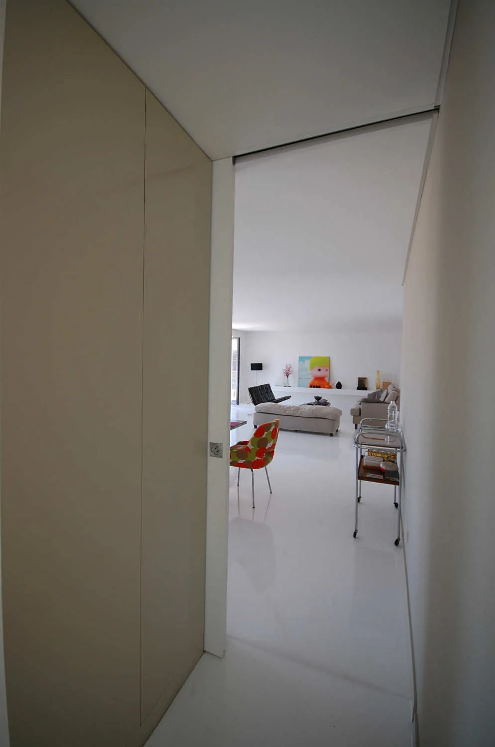 A view from a hallway into a minimalist white living room featuring glossy reflective floors, a patterned accent chair, and a long art ledge.