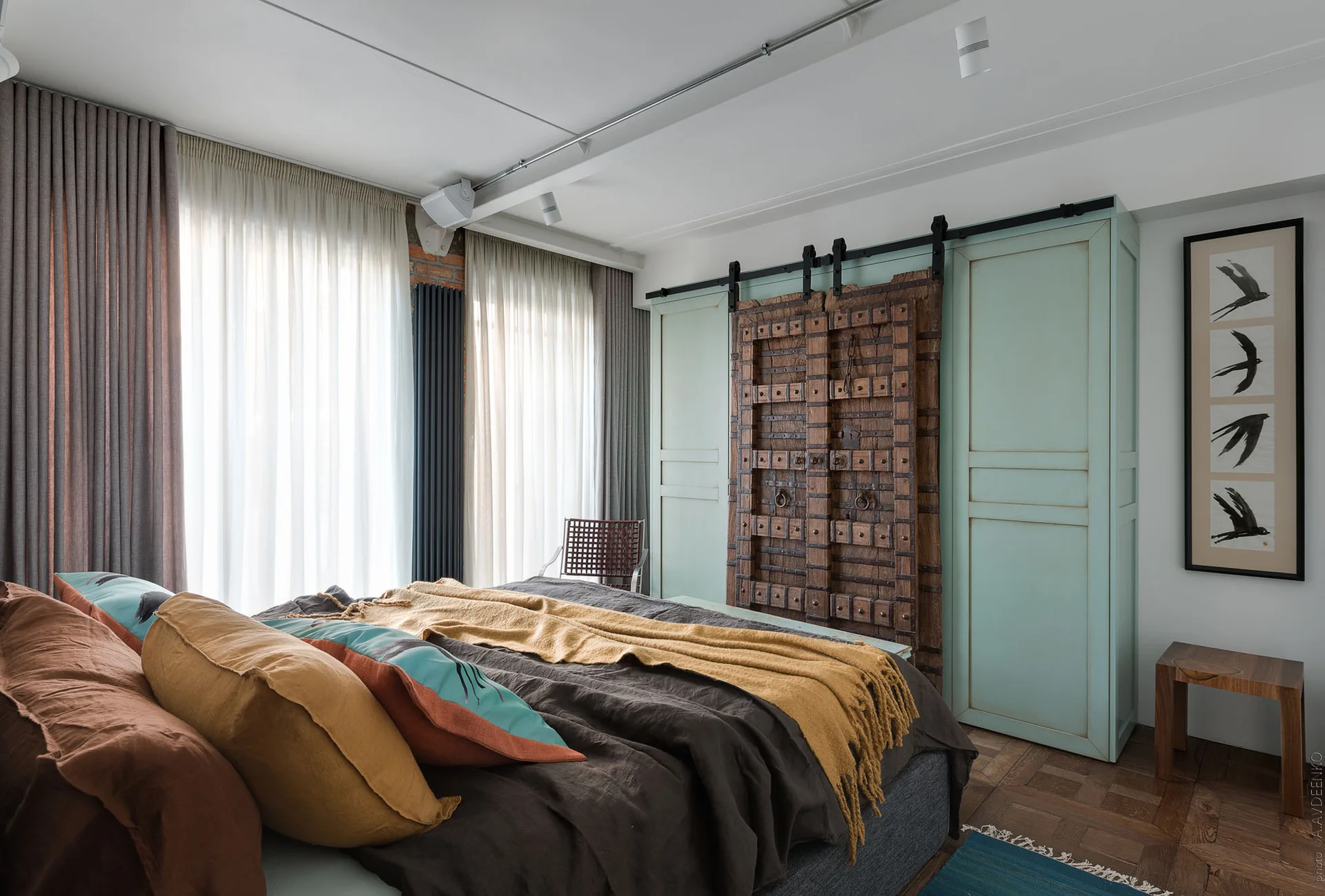 A bedroom features a bed with earthy linens, parquet flooring, and a pale green wardrobe with an antique wooden sliding barn door.