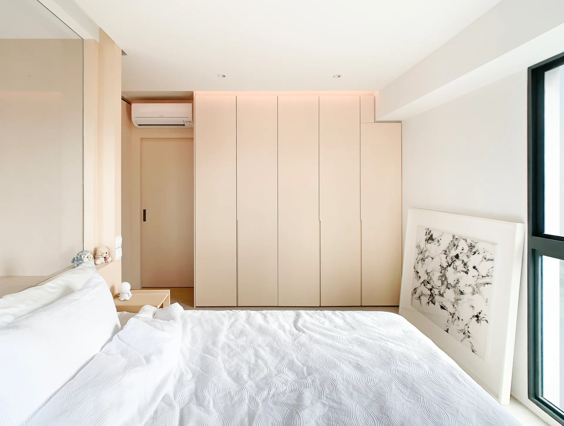 A minimalist bedroom featuring a bed with textured white sheets next to a large built-in beige wardrobe and a framed black-and-white abstract painting.