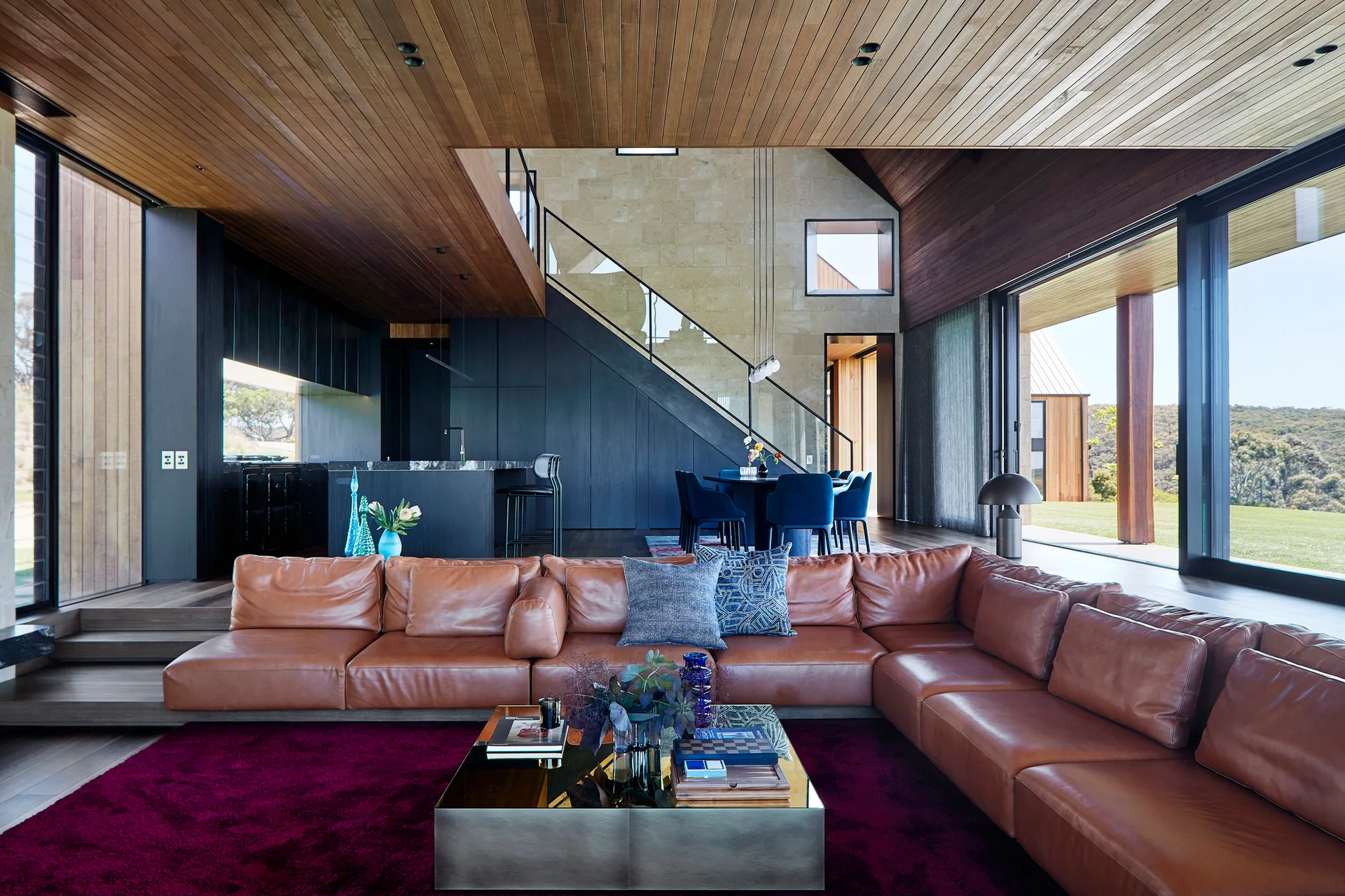 A tan leather sectional sits on a deep magenta rug in a living room with a wood ceiling, a stone accent wall, and glass sliding doors.