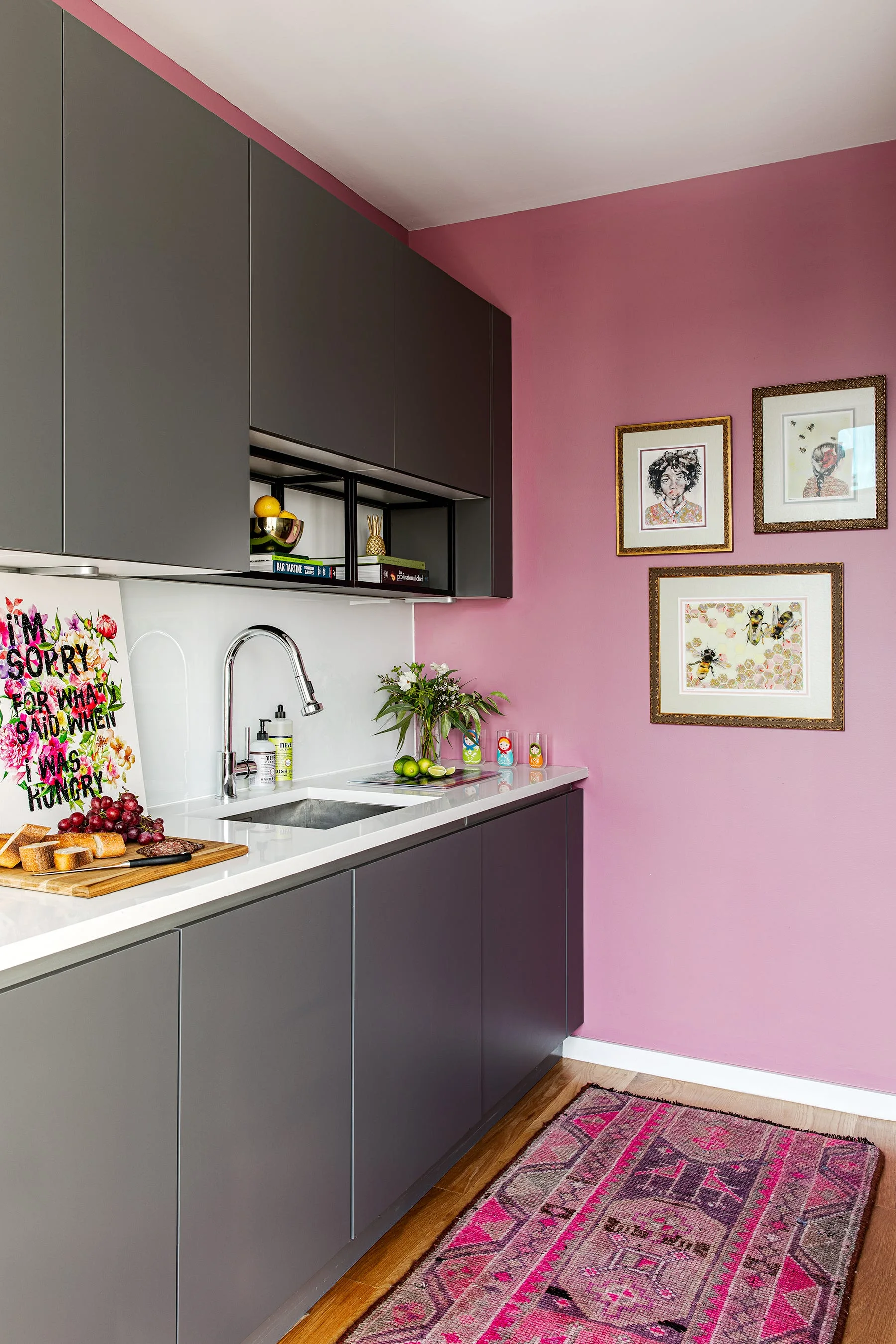 A modern kitchenette featuring flat-panel dark grey cabinets, a white countertop, a vibrant pink wall with art, and a patterned rug.