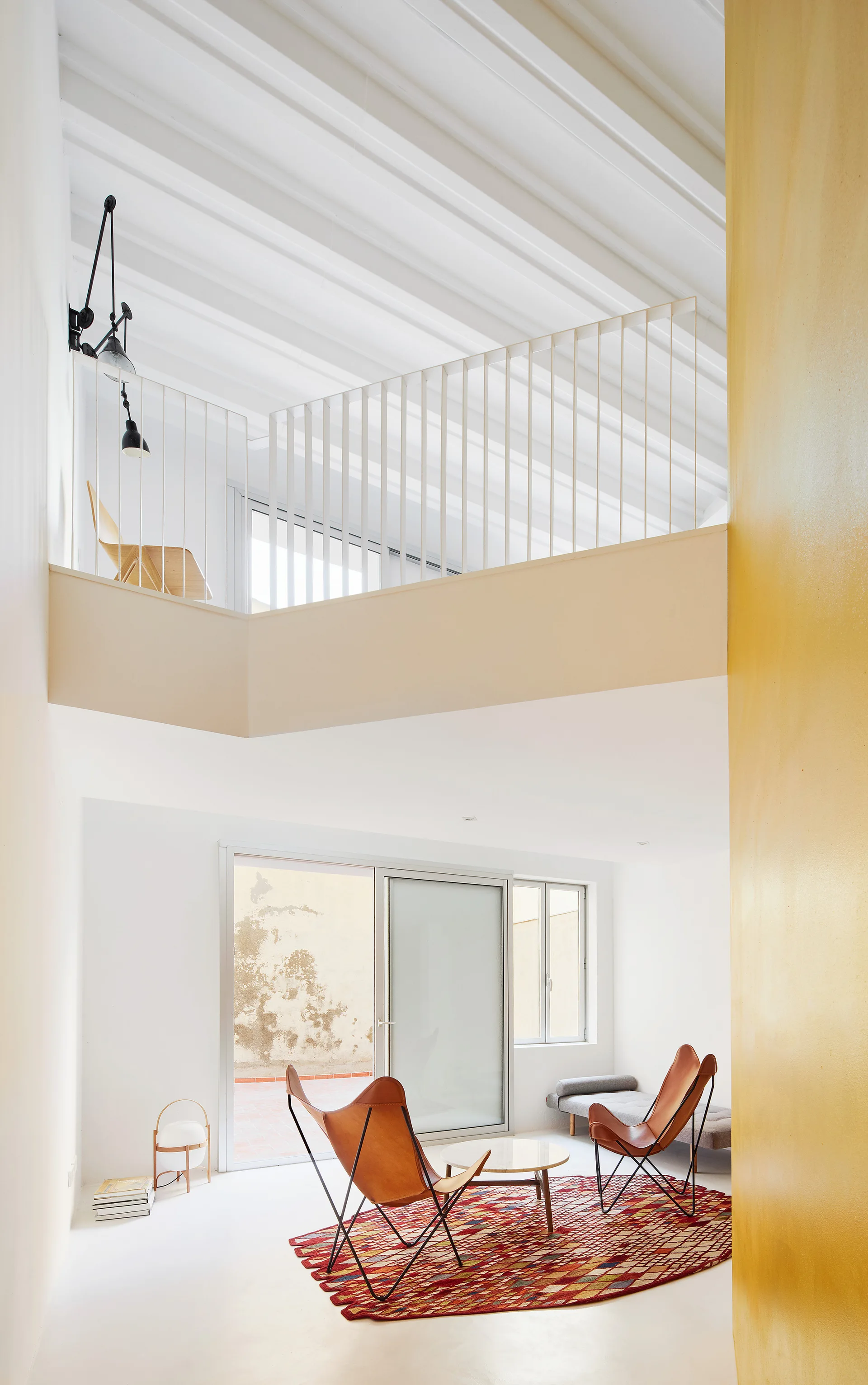 A bright white double-height living room featuring two leather butterfly chairs on a red patterned rug beneath a mezzanine loft.