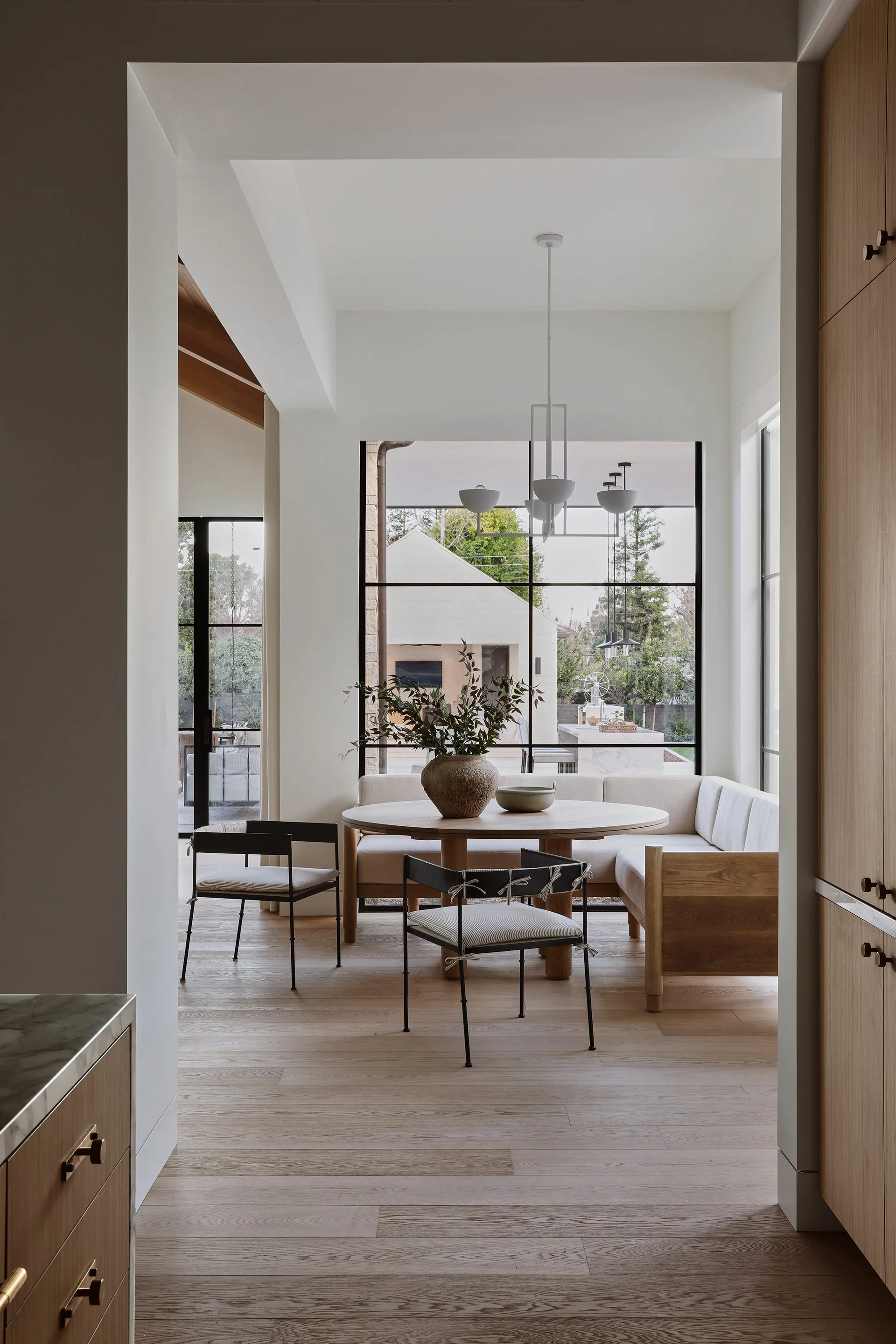 A light-filled dining nook with a circular wood table, built-in corner banquette, and black-framed chairs against a large grid window.
