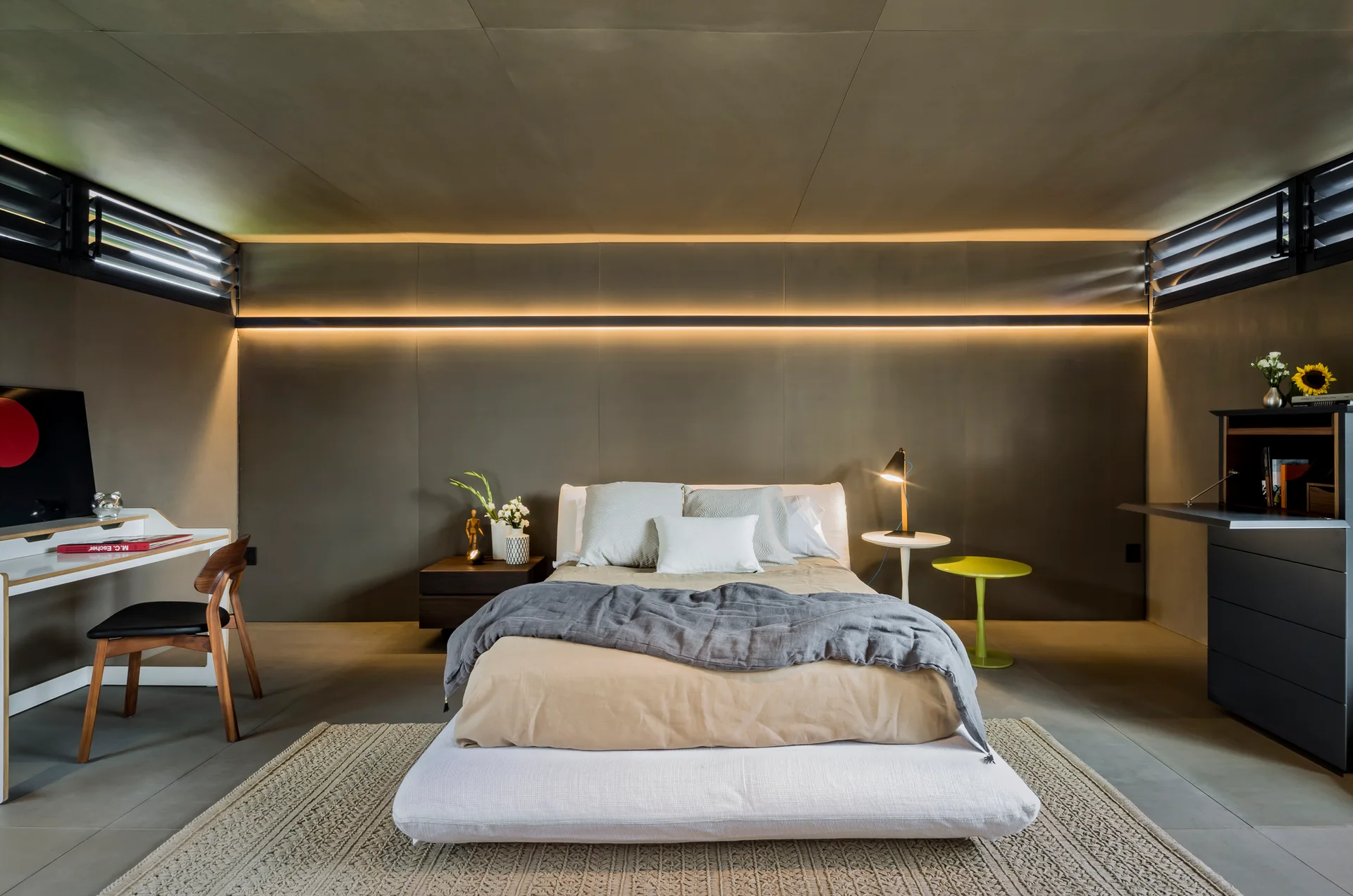 A modern bedroom features a low platform bed, a white desk, a dark storage cabinet, and a horizontal strip of warm light on the paneled back wall.