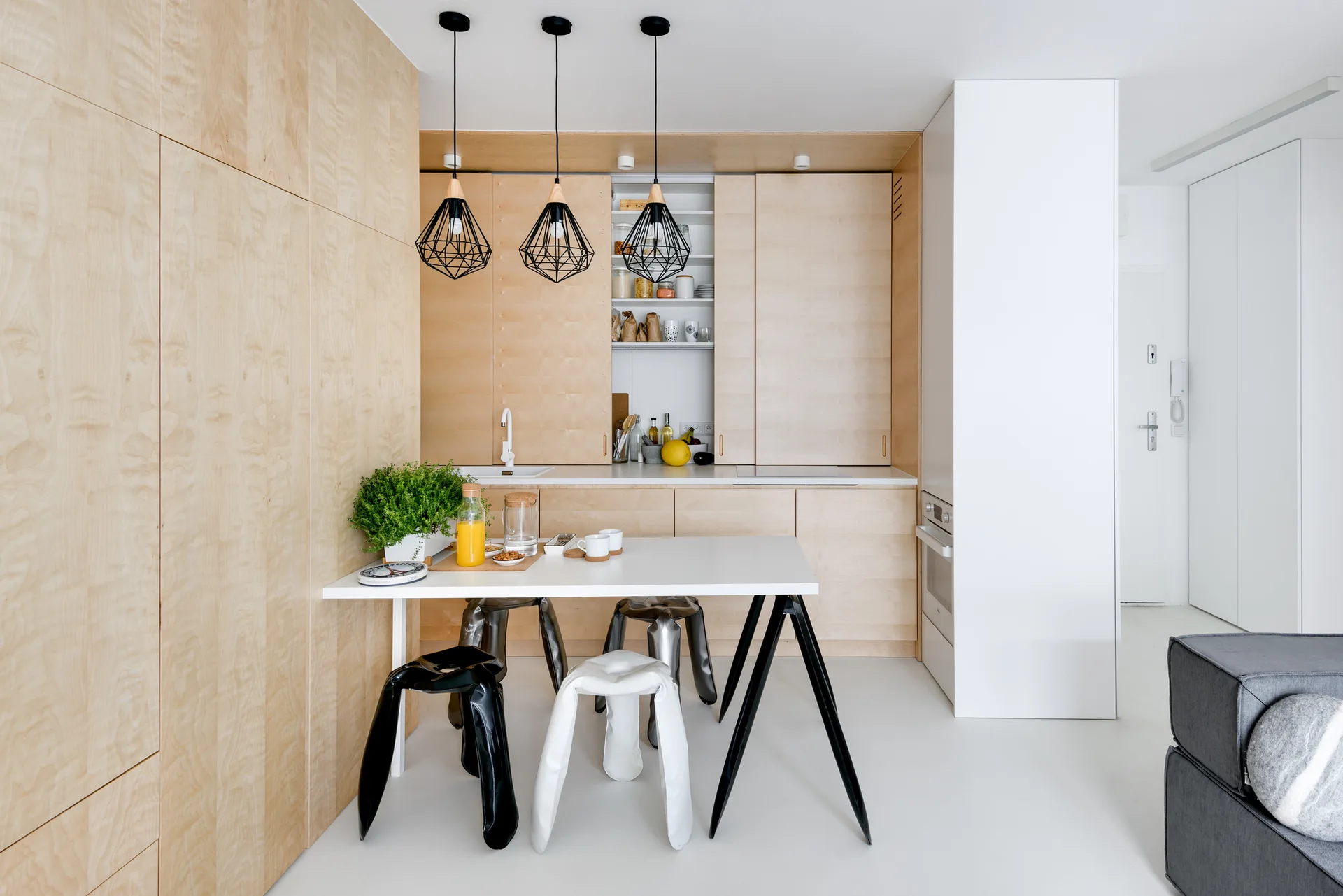 A minimalist kitchen features light wood paneled cabinetry, a white dining table with black and white sculptural stools, and geometric pendant lights.