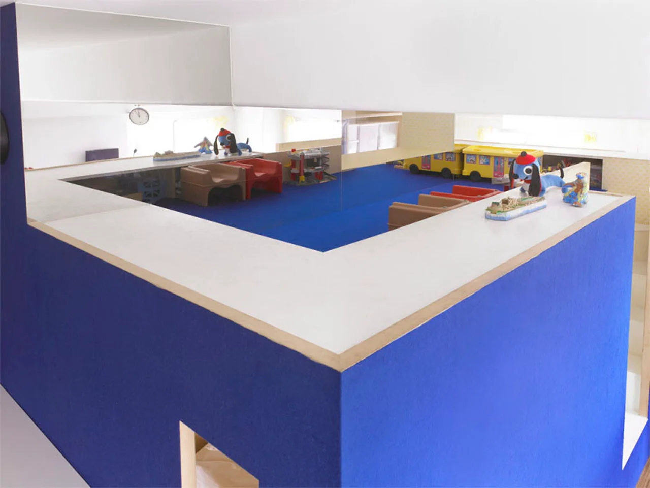 A children's play loft features a raised cobalt blue platform with a white ledge, small plastic armchairs, and a large horizontal wall mirror.