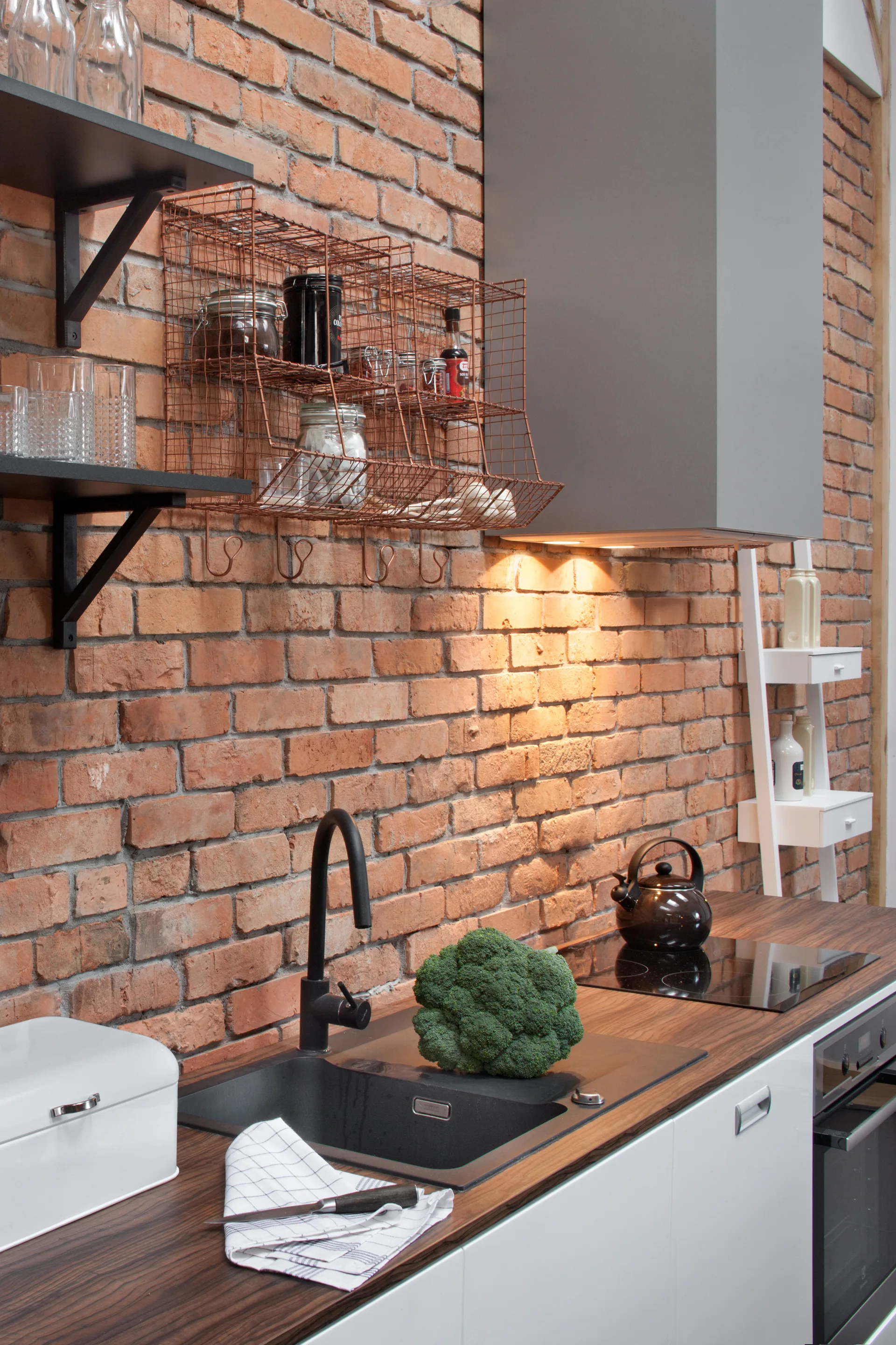 A modern kitchen features an exposed red brick wall, a dark wood-toned countertop with a black undermount sink, and a copper wire organizer.