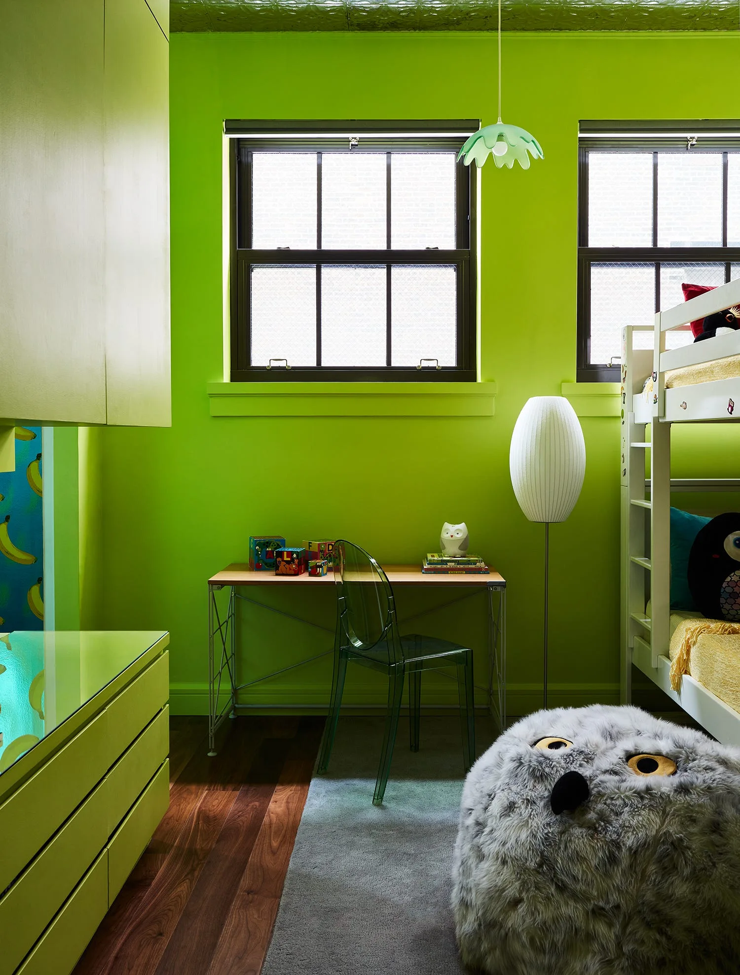 A kids bedroom features bright lime green walls, a wooden desk with a transparent chair, a white bunk bed, and a large furry owl pouf.