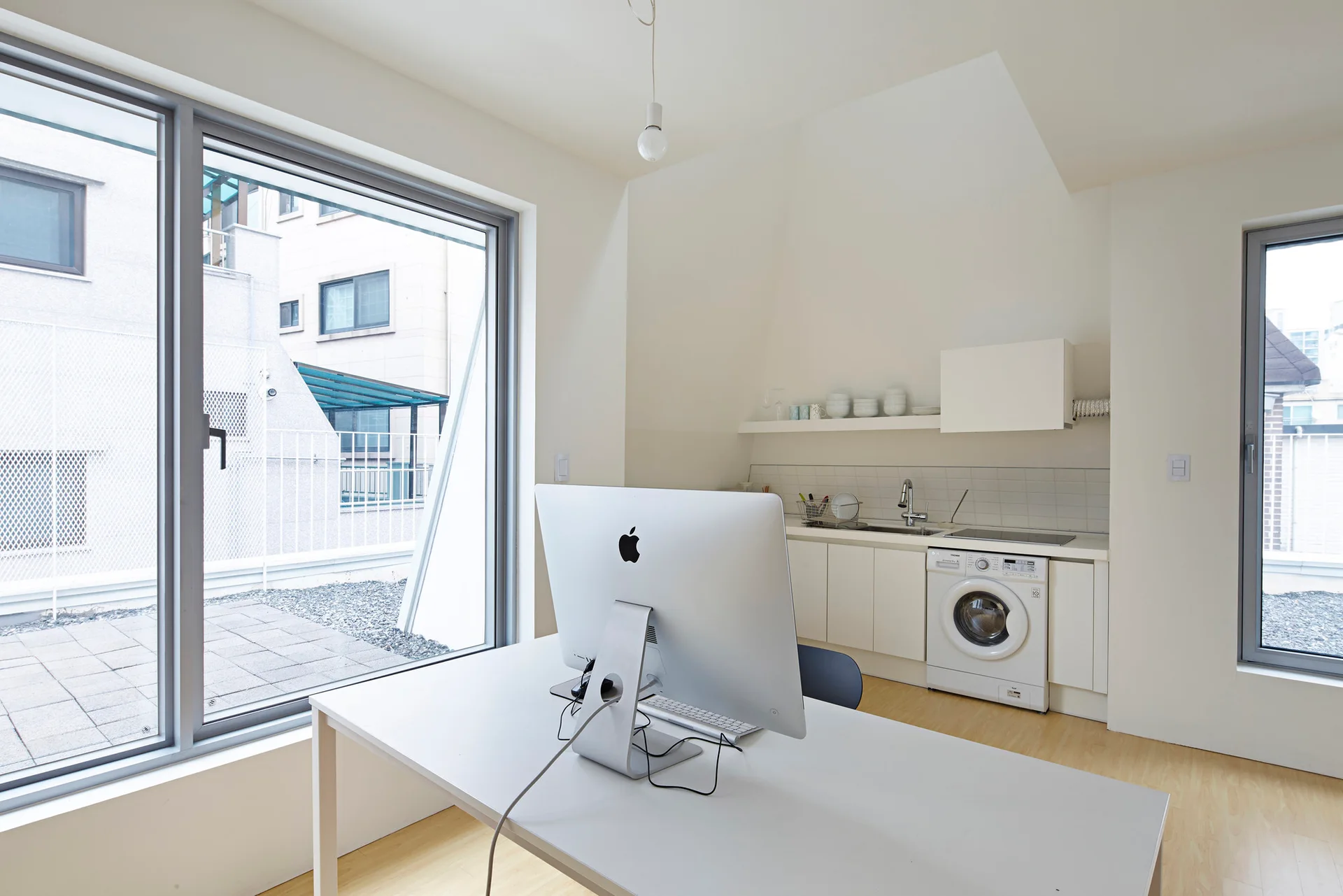 A white desk with a desktop computer sits in a minimalist office next to a compact kitchenette featuring a built-in washing machine.