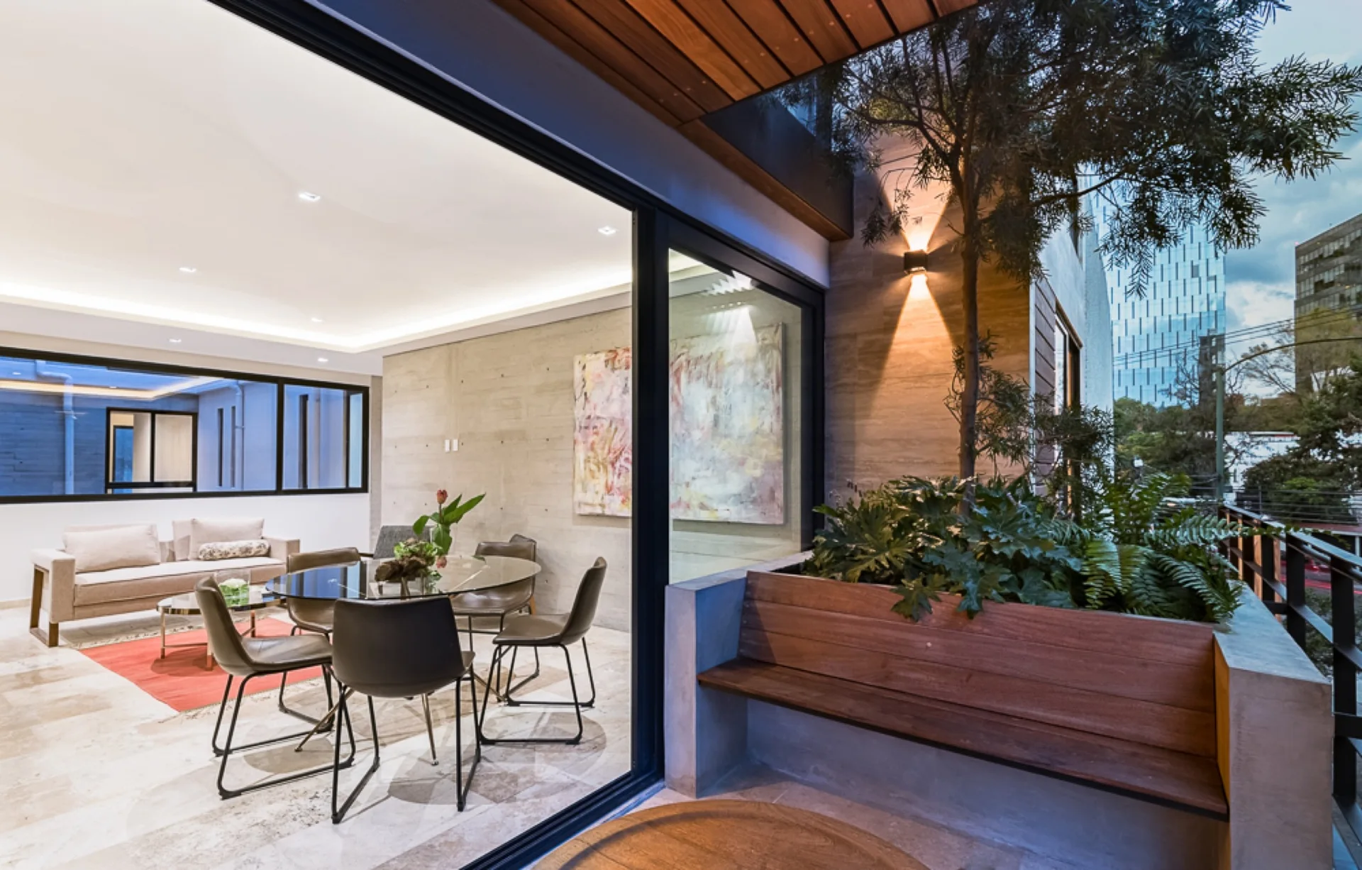 A modern dining room with a glass table and concrete walls opens through sliding glass doors to a terrace featuring a built-in wood and concrete bench.