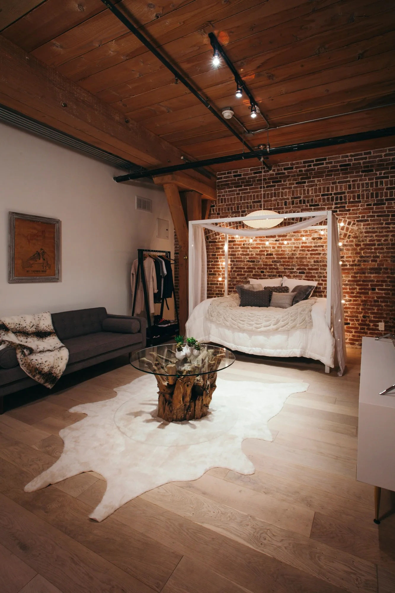 An industrial loft bedroom features a white canopy bed against an exposed brick wall, a grey sofa, and a glass-top driftwood coffee table.