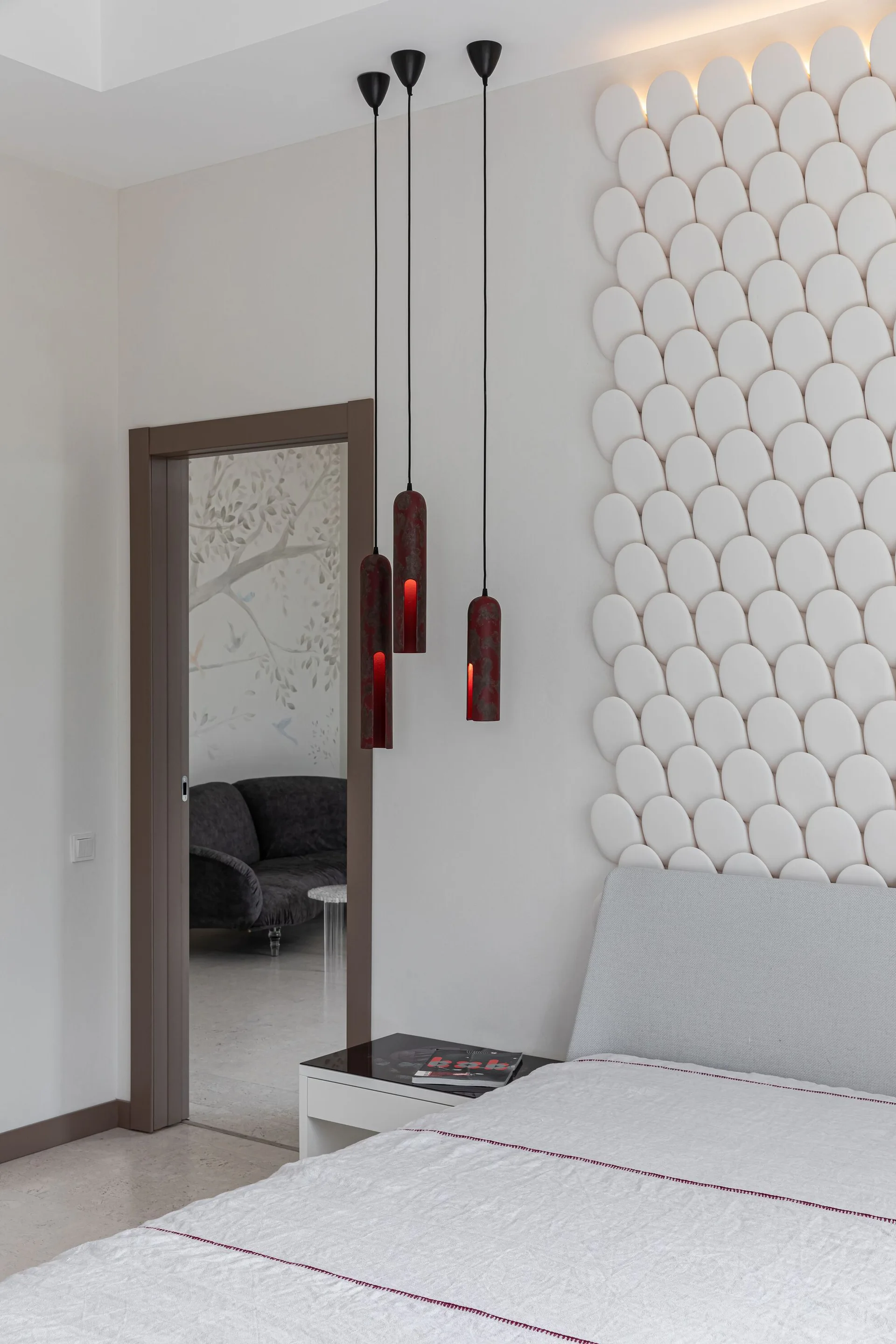 A modern bedroom featuring a bed with a light grey headboard, a textured white oval wall installation, and three hanging red pendant lights.