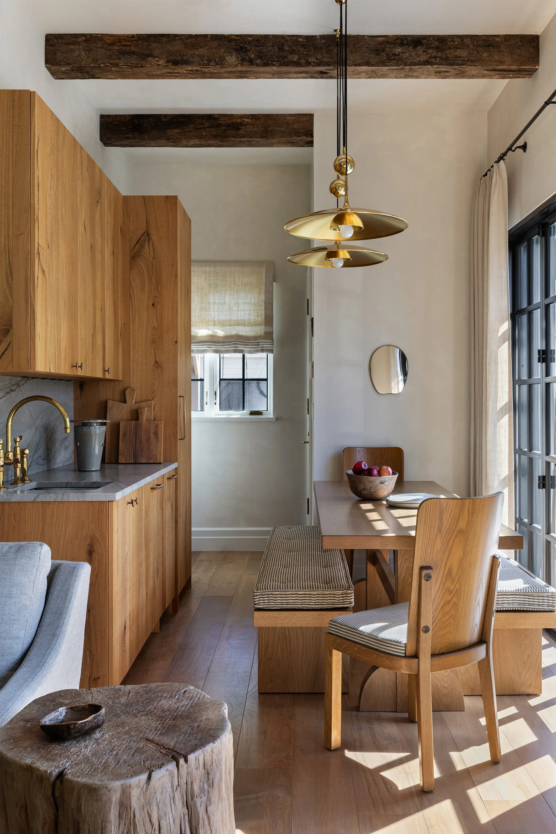 A wooden dining table with striped bench cushions sits next to a light wood kitchenette beneath dark rustic ceiling beams.