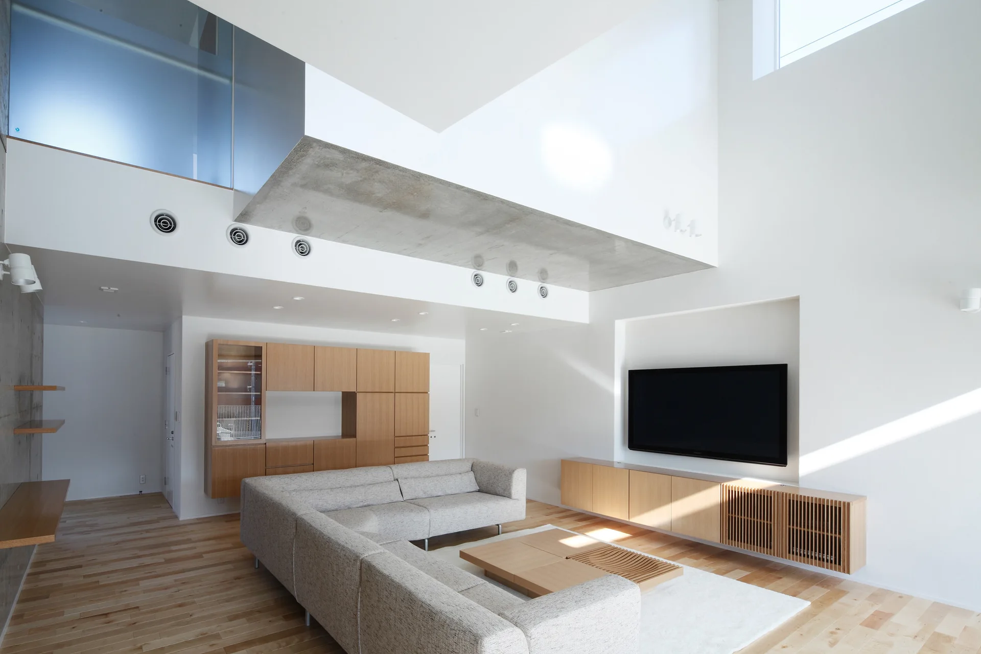 A minimalist living room features a grey sectional sofa, light wood cabinets, and an exposed concrete ceiling beam.