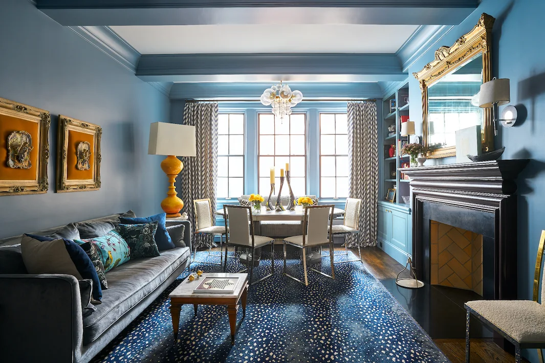 An eclectic living and dining area features deep blue walls, a gray velvet sofa, a fireplace with a gold mirror, and a dining table by the window.
