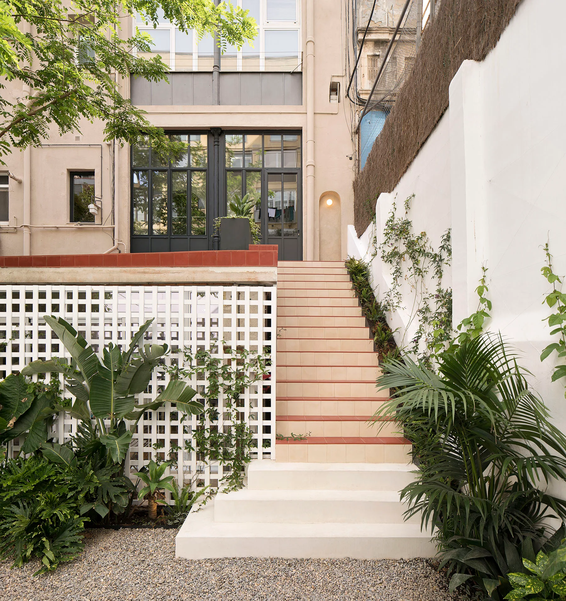 A landscaped outdoor patio features a central tiled staircase flanked by green plants, a white grid trellis, and gravel ground cover.
