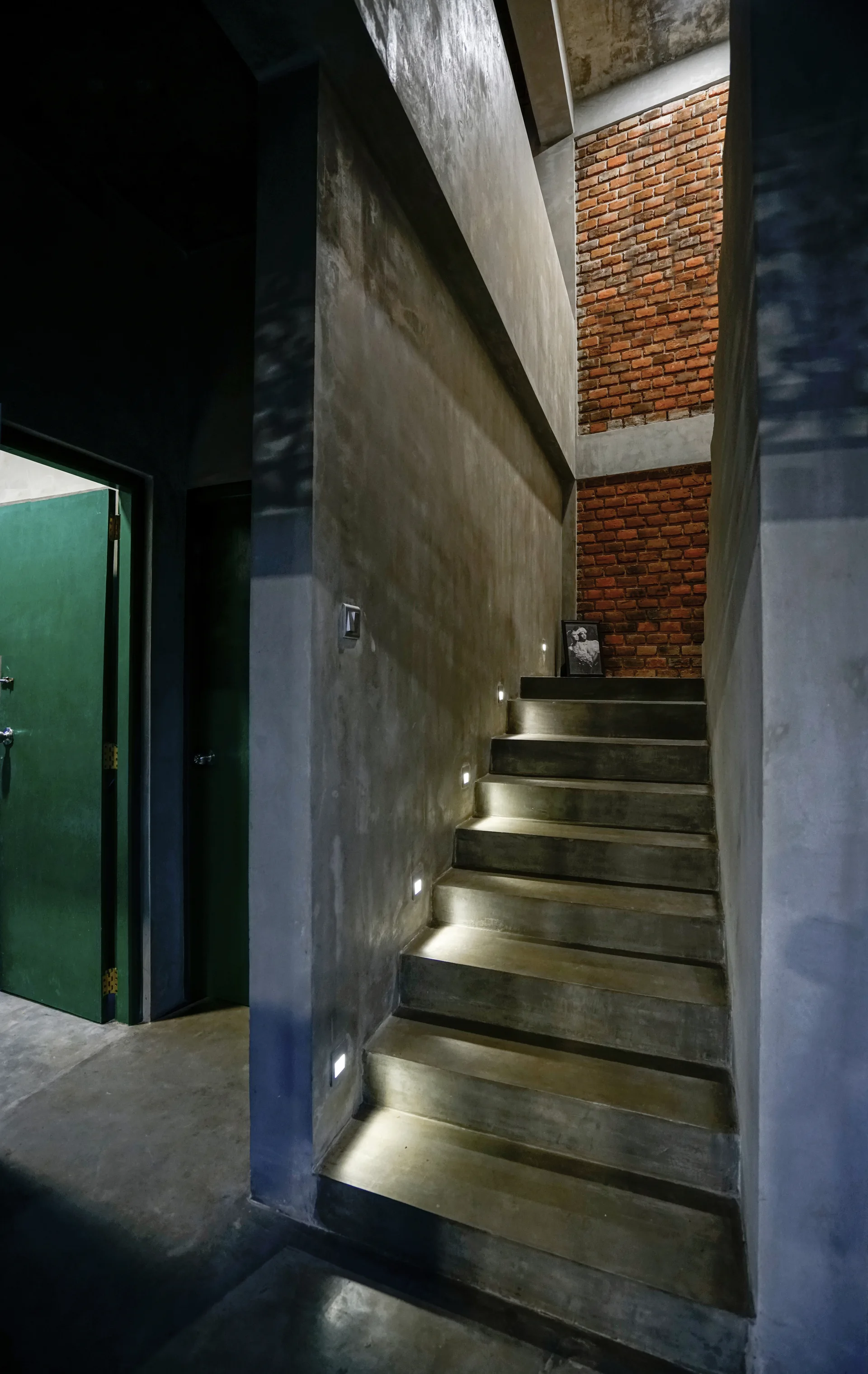 A concrete staircase with integrated step lighting stands next to a textured red brick wall and a green door.